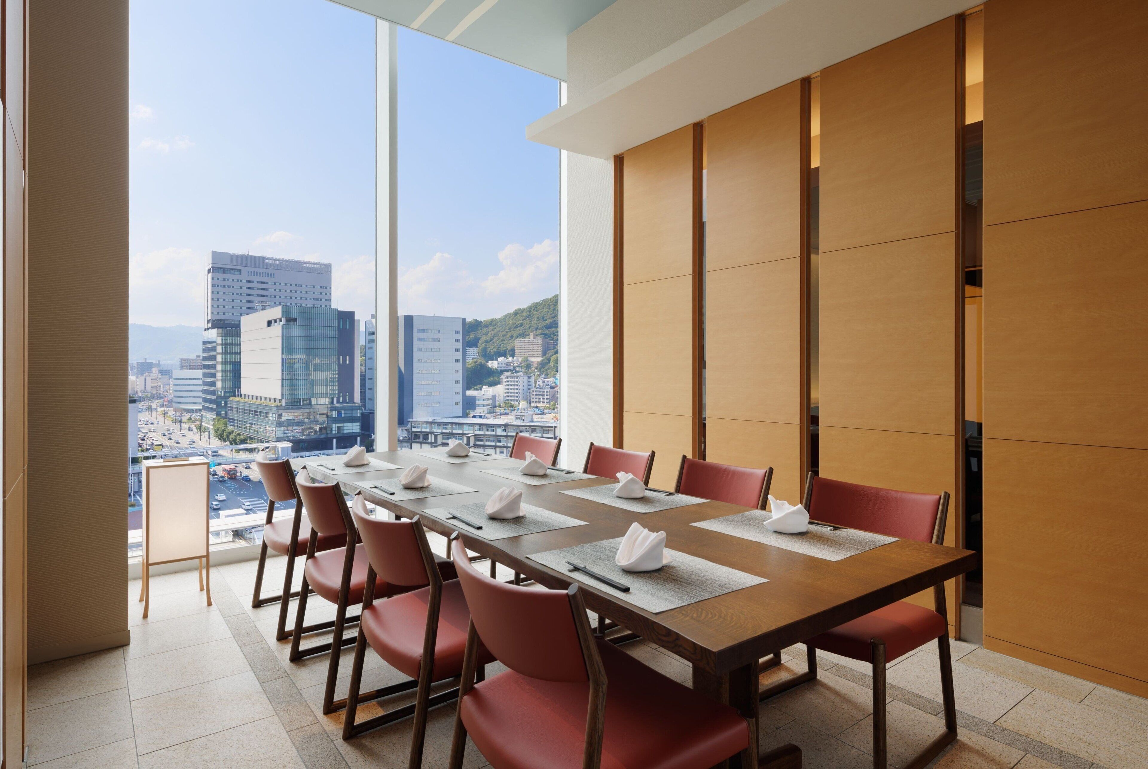 A conference room with a long table, red chairs, and a city view through large windows.