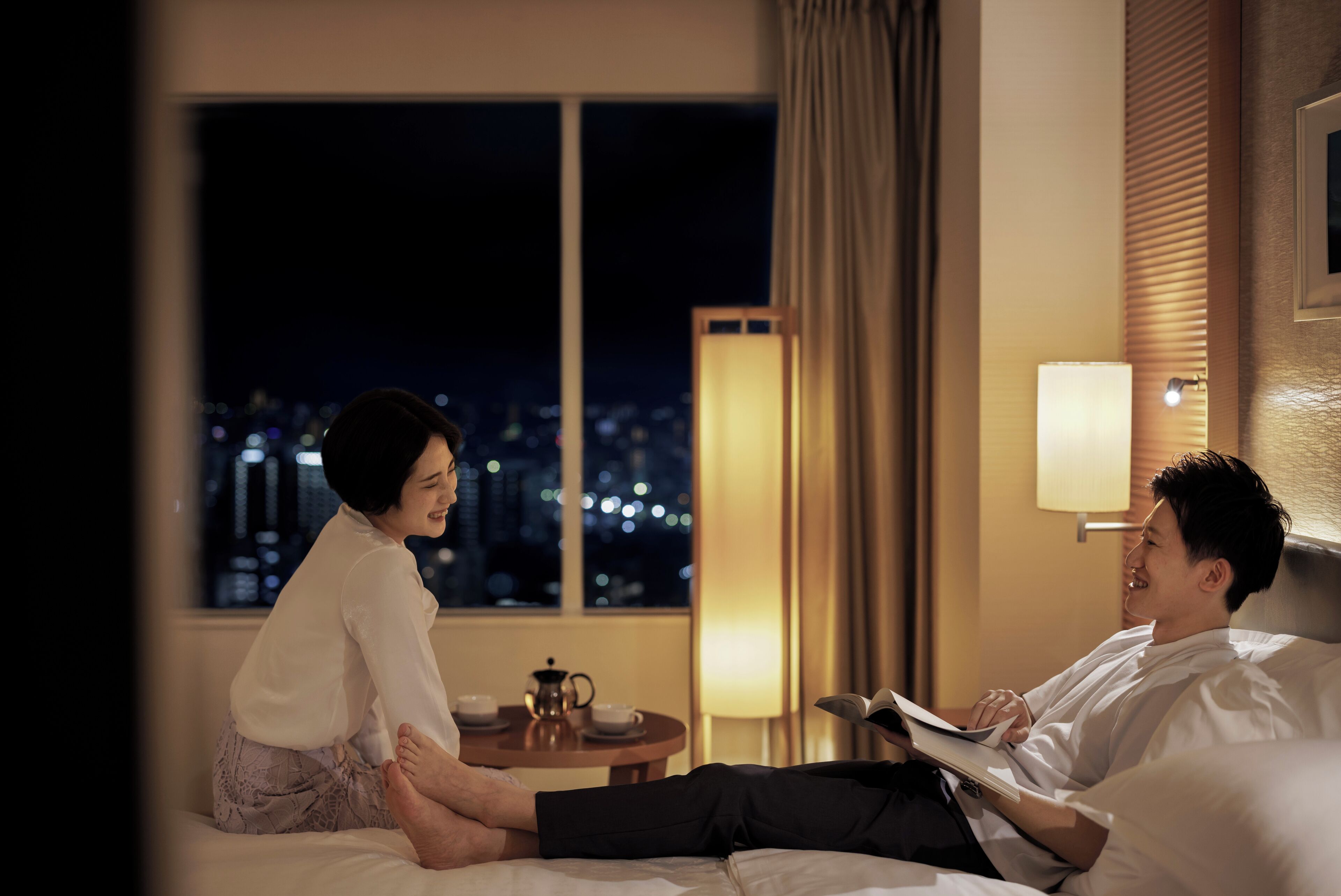 A hotel room with a bed, a bedside table with a lamp, a window with a view of the city at night, and a cityscape visible through the window.
