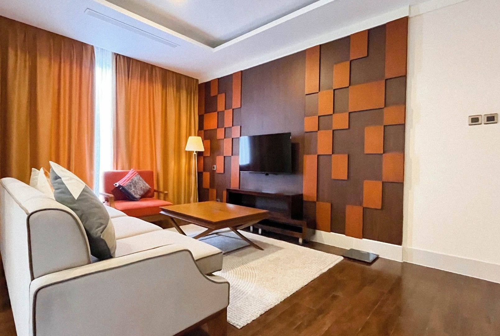A modern living room with a sofa, a red armchair, a coffee table, a flat-screen TV mounted on a geometric-patterned wall, and a floor lamp.