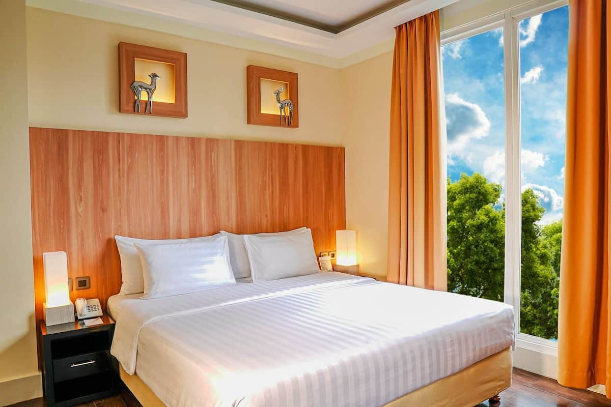A hotel room with a large bed, wooden headboard, two wall-mounted picture frames, a nightstand with a lamp, and a view of trees through a window.
