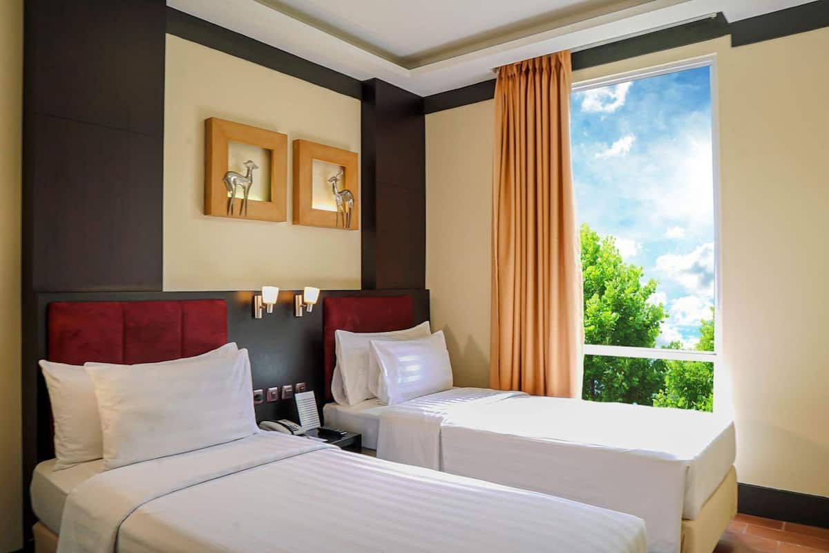 A hotel room with two beds, a large window with a view of greenery, and wall-mounted light fixtures.