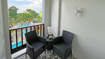 A balcony with two wicker chairs, a small table, and a potted orchid. A pool is visible through the balcony, surrounded by greenery and buildings.
