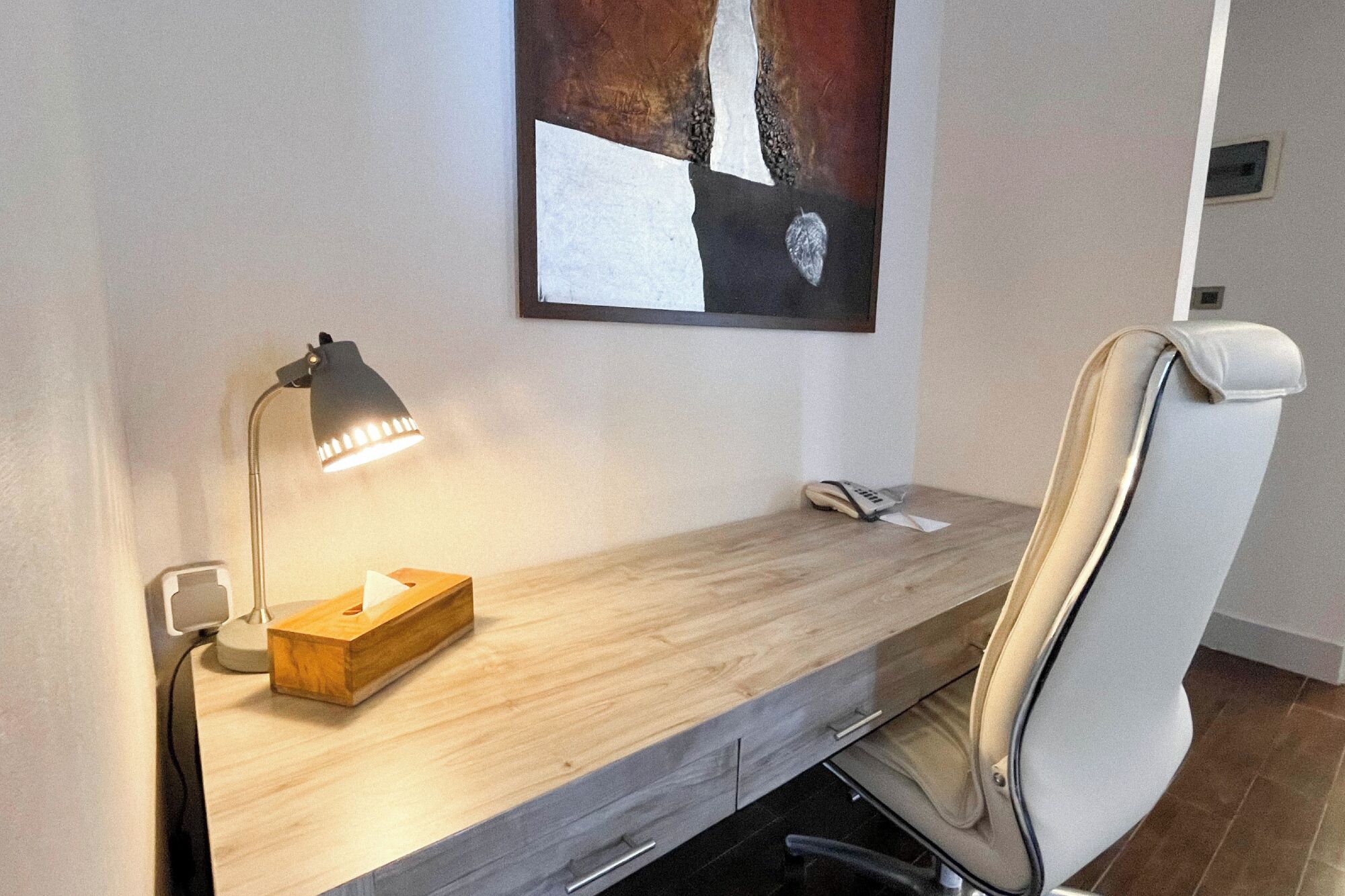 A modern office chair with a high backrest, a desk with a wooden surface, a desk lamp, and a tissue box.