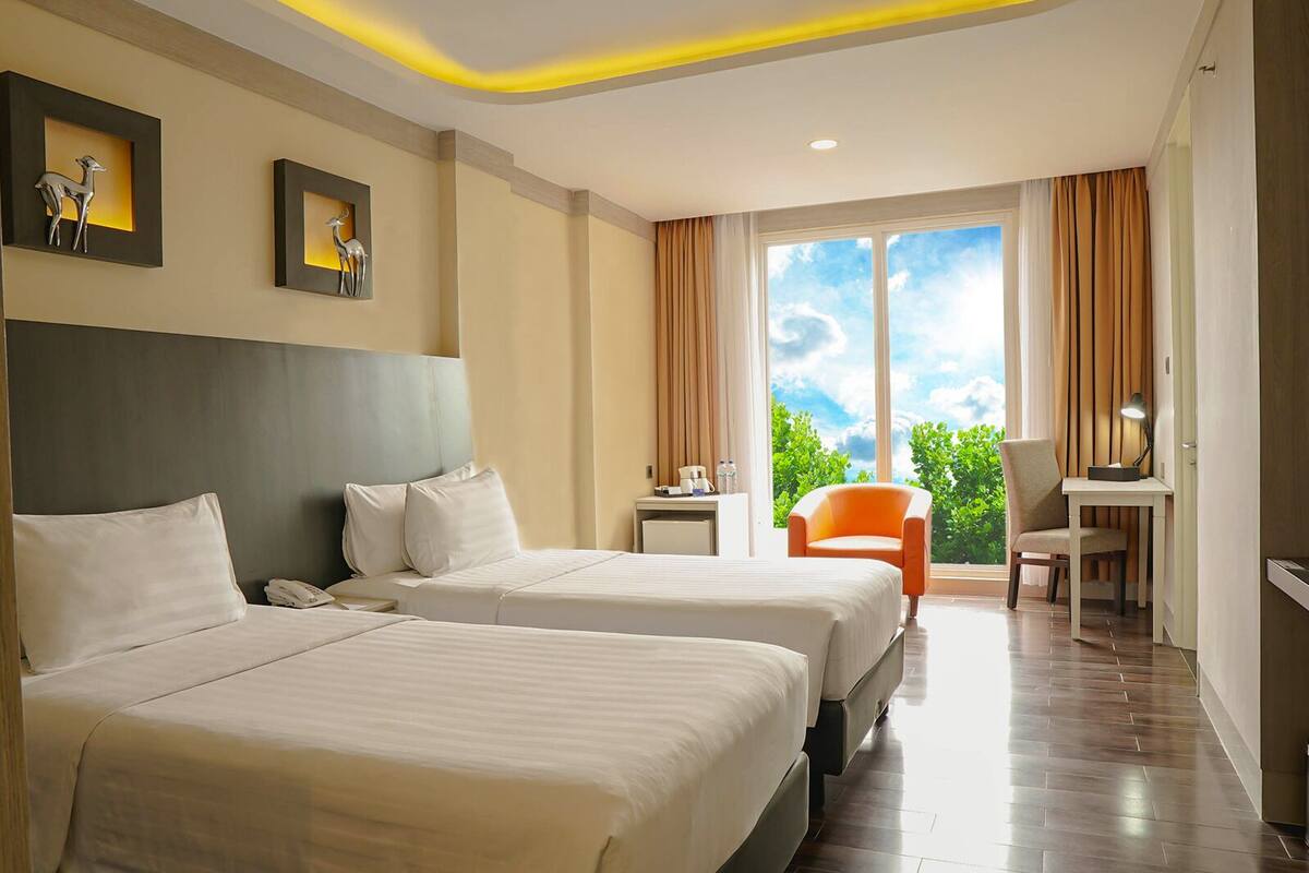 A hotel room with two beds, a desk, a chair, and a view of greenery through the window.