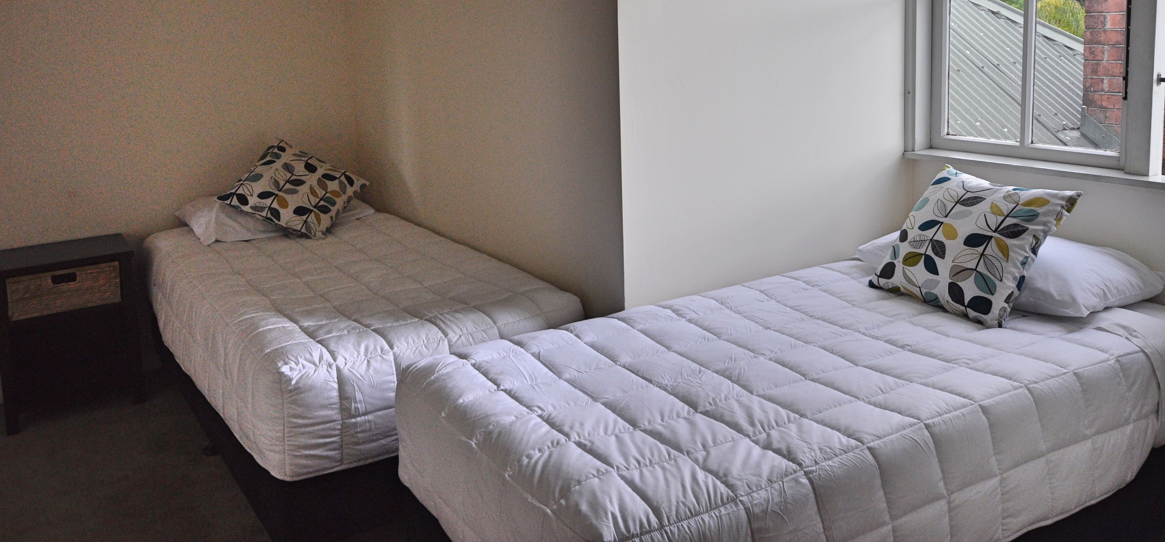 Two single beds with white bedding and patterned pillows, a wooden nightstand, and a window with a view of a building.