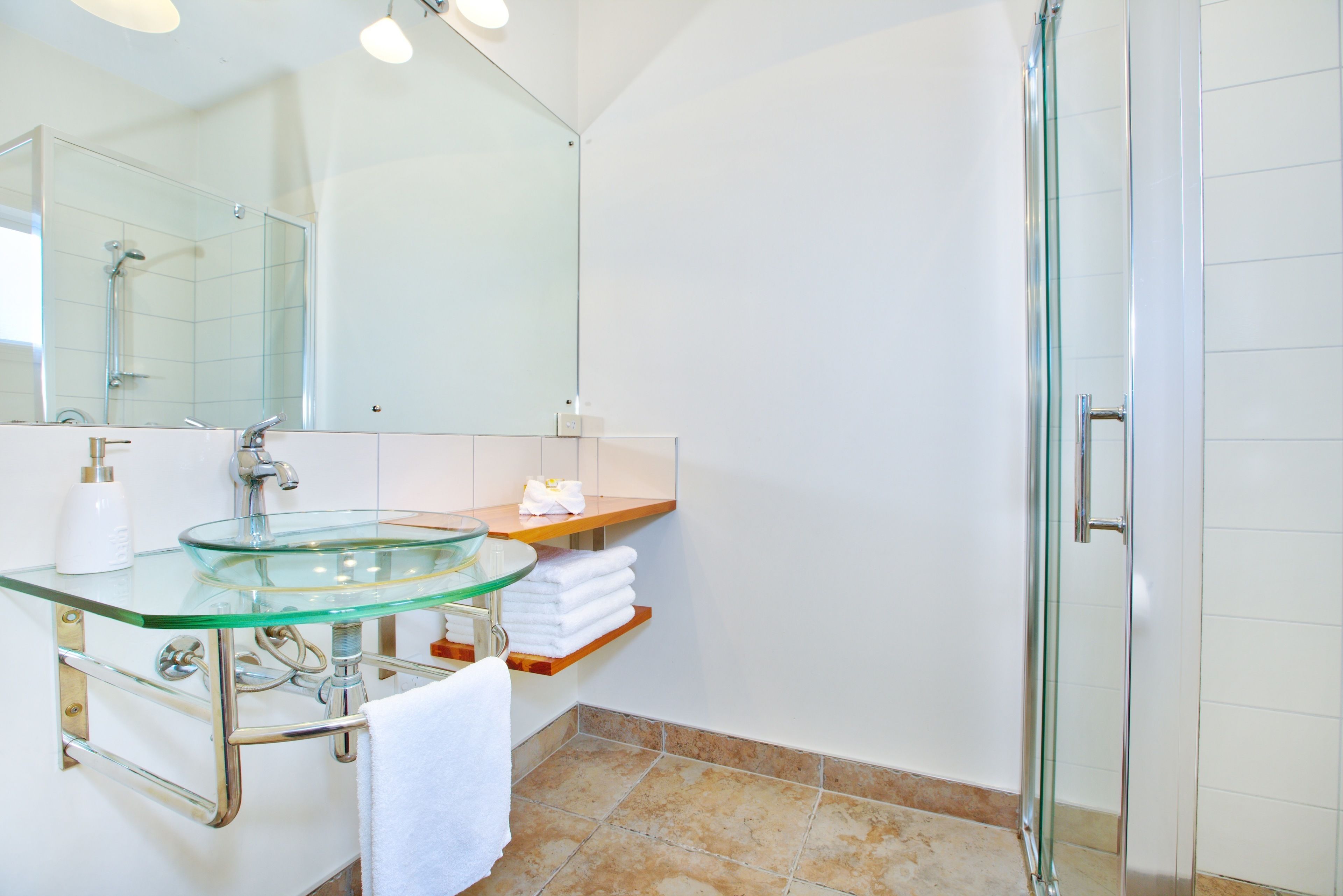 A modern bathroom with a glass sink, a large mirror, a glass shower enclosure, and a white tiled floor.