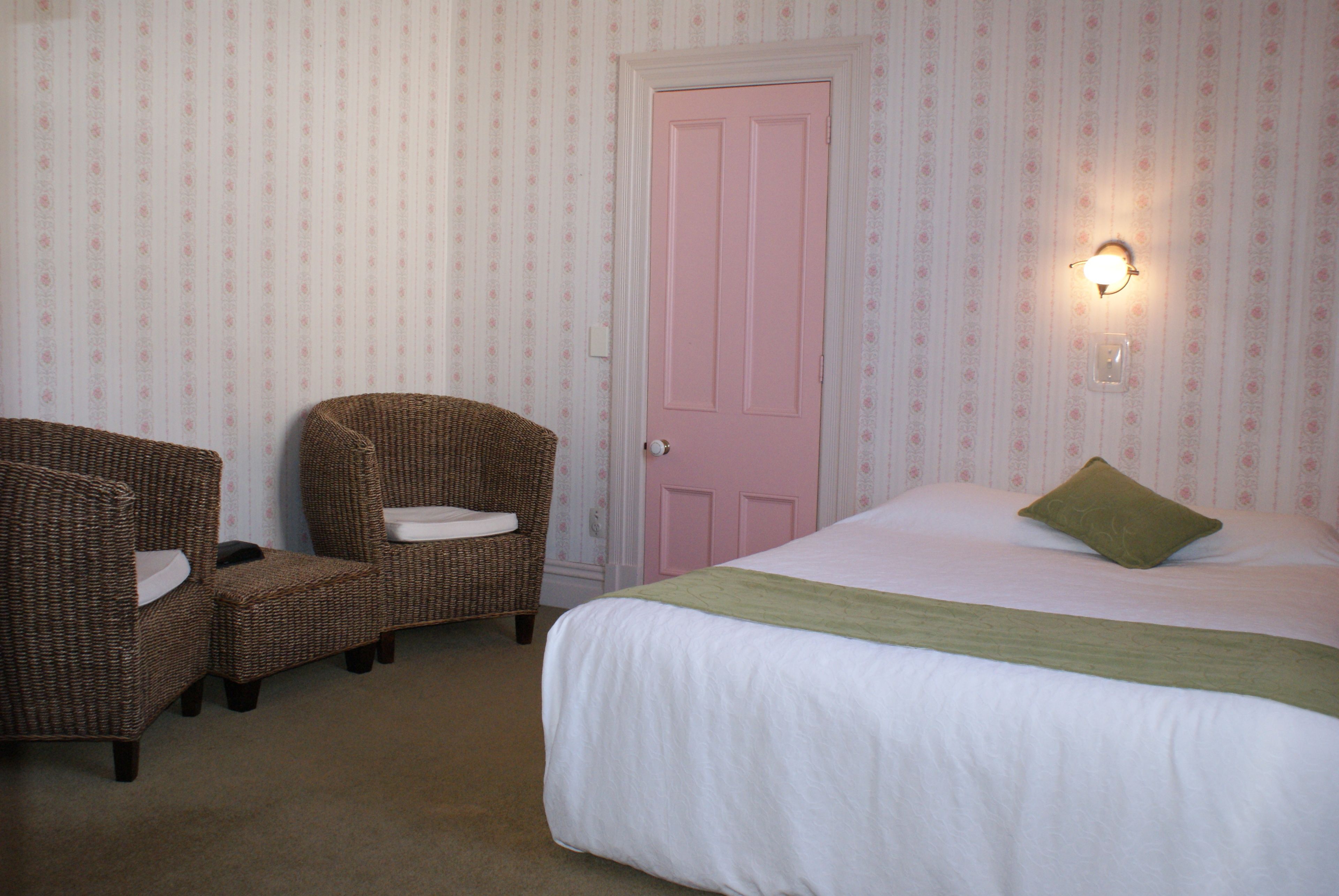 A bedroom with a bed, two wicker chairs, a pink door, and a wall-mounted light.