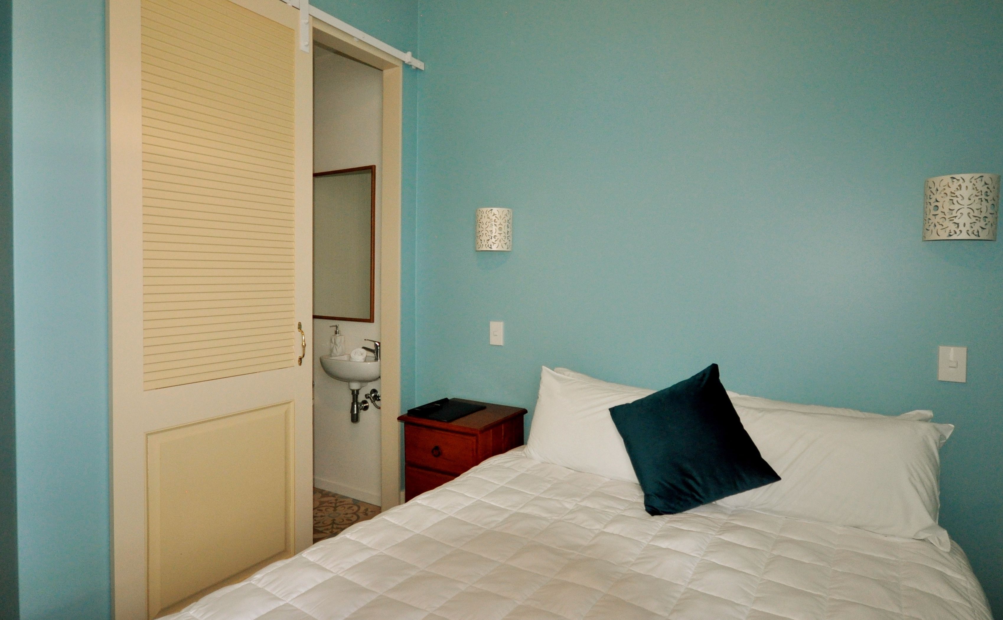 Un dormitorio con cama, mesita de noche, cómoda, puerta, espejo y lámpara fijada en la pared.