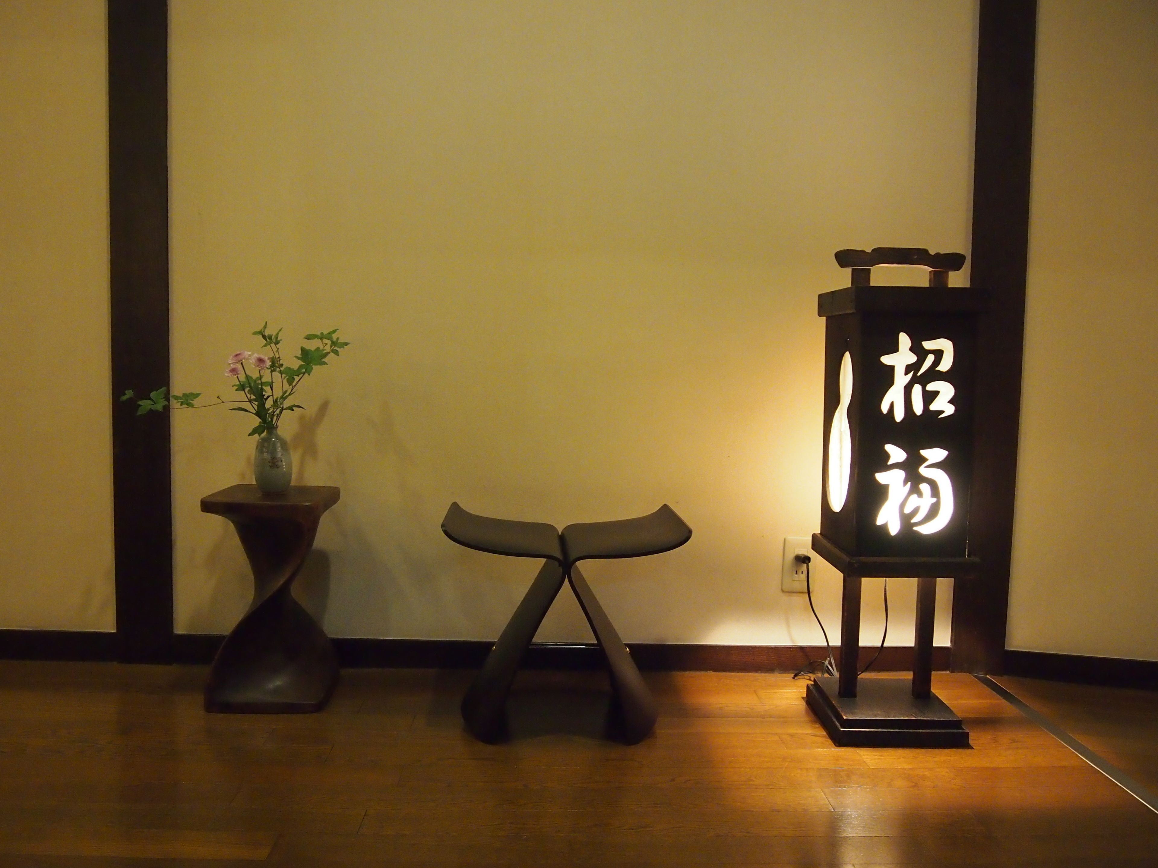 A room with a wooden floor, a dark-colored table with a vase of flowers, and a stand with illuminated calligraphy.