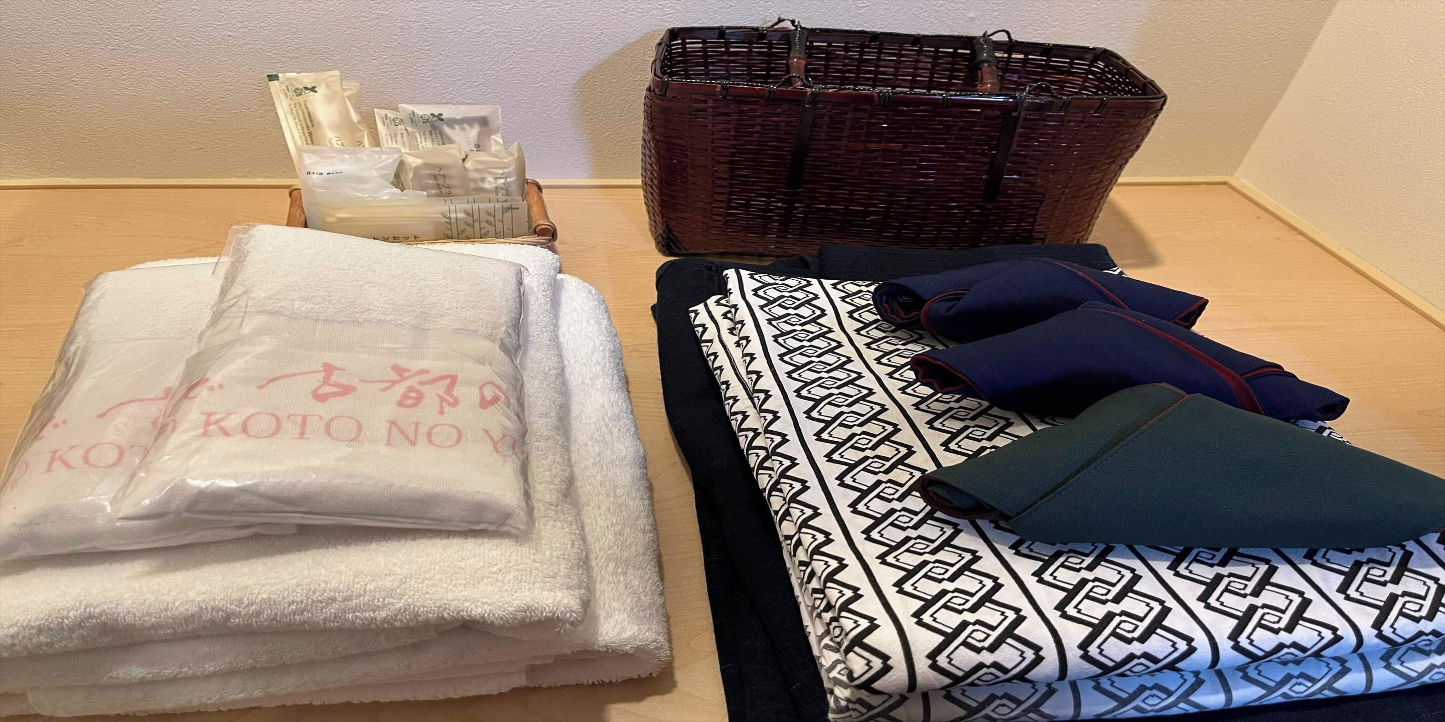Une pièce bien rangée, avec une pile de serviettes blanches, une couverture à motifs, un coussin bleu foncé et vert, et un panier en osier contenant des articles de toilette.