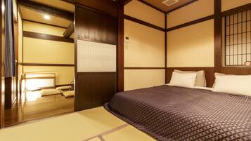 A traditional Japanese room with tatami flooring, a low wooden bed with a patterned bedspread, and a sliding door leading to a washbasin area.