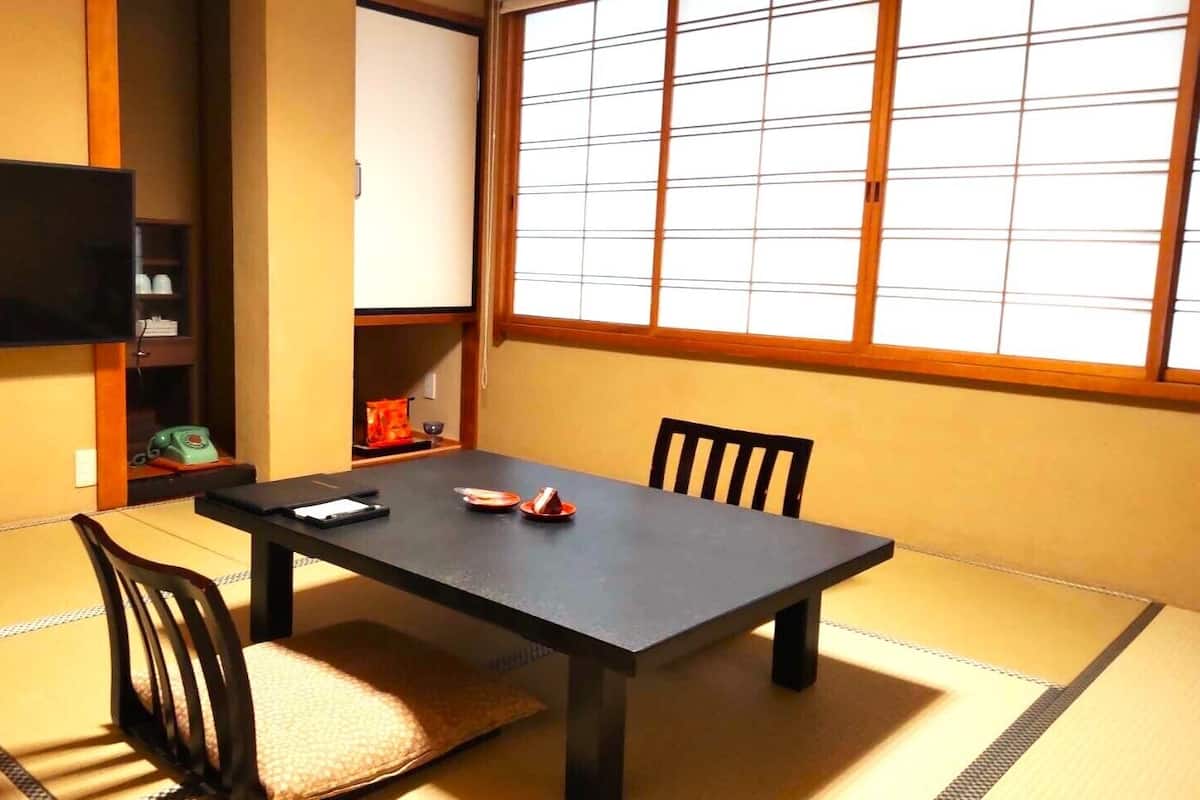 Una habitación japonesa tradicional con suelo de tatami, una mesa baja, dos sillas, un televisor y una puerta corredera.