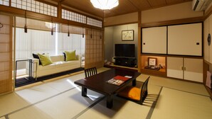Una habitación tradicional japonesa con suelo de tatami, una mesa baja, un sofá con cojines, un televisor y puertas correderas.