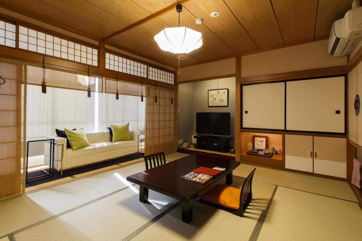 Una habitación tradicional japonesa con suelo de tatami, una mesa baja, un sofá con cojines, un televisor y puertas correderas.