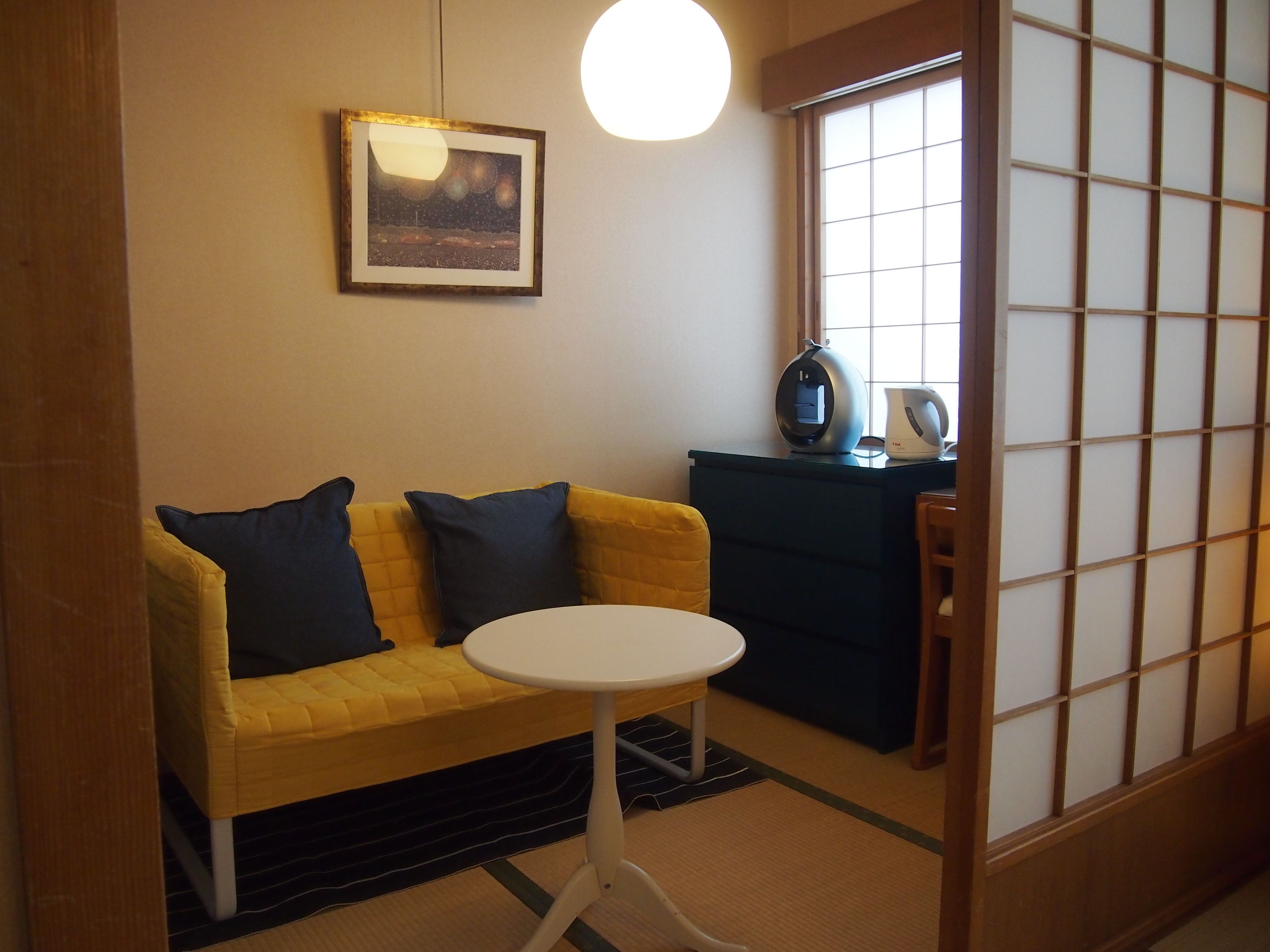 Una habitación con un sofá amarillo, una mesita blanca pequeña, una puerta corredera tradicional japonesa y un cuadro en la pared.