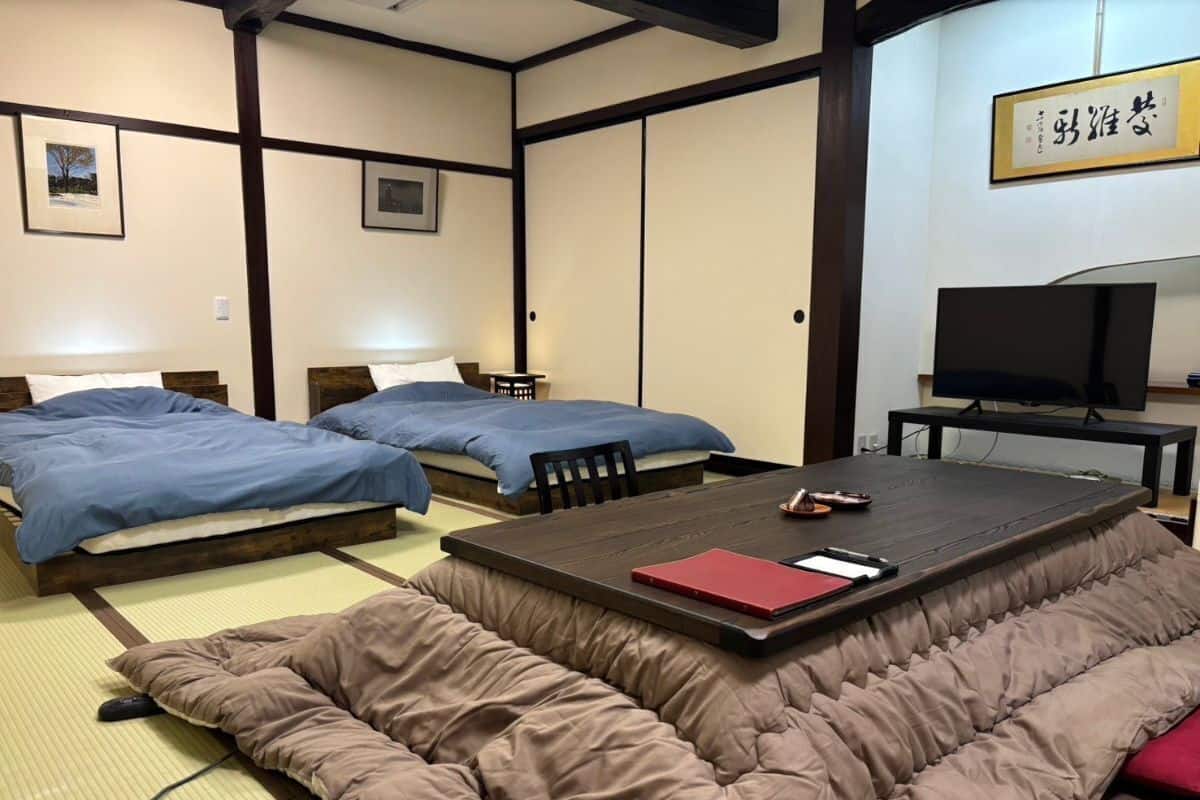 Una habitación tradicional japonesa con suelo de tatami, dos camas con ropa de cama azul, un sofá de tatami, un televisor y una obra de caligrafía enmarcada en la pared.