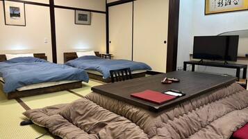 A traditional Japanese-style room with tatami flooring, two beds with blue bedding, a tatami mat sofa, a television, and a framed calligraphy piece on the wall.