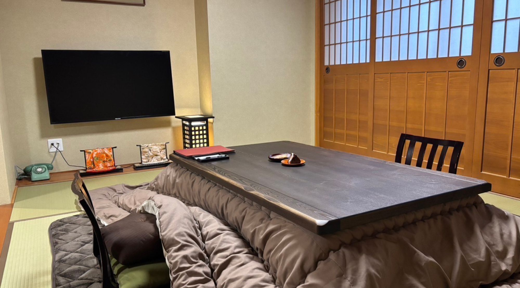 A traditional Japanese-style room with tatami flooring, a low table, cushions, a television, and a sliding door with wooden panels.