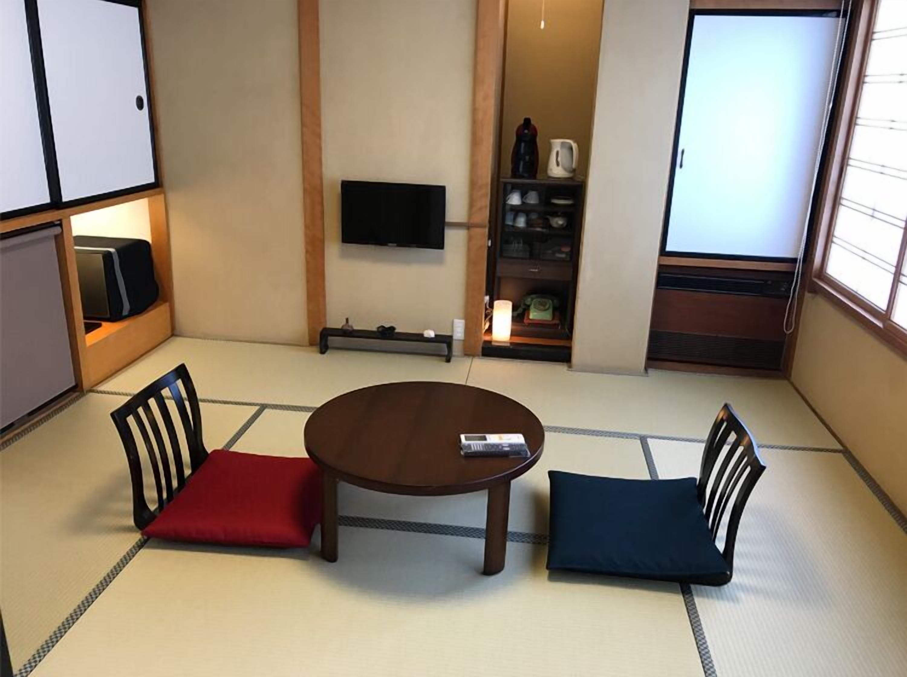 Una habitación con tatamis, una mesa baja, dos sillas, un televisor y una estantería con utensilios de cocina.