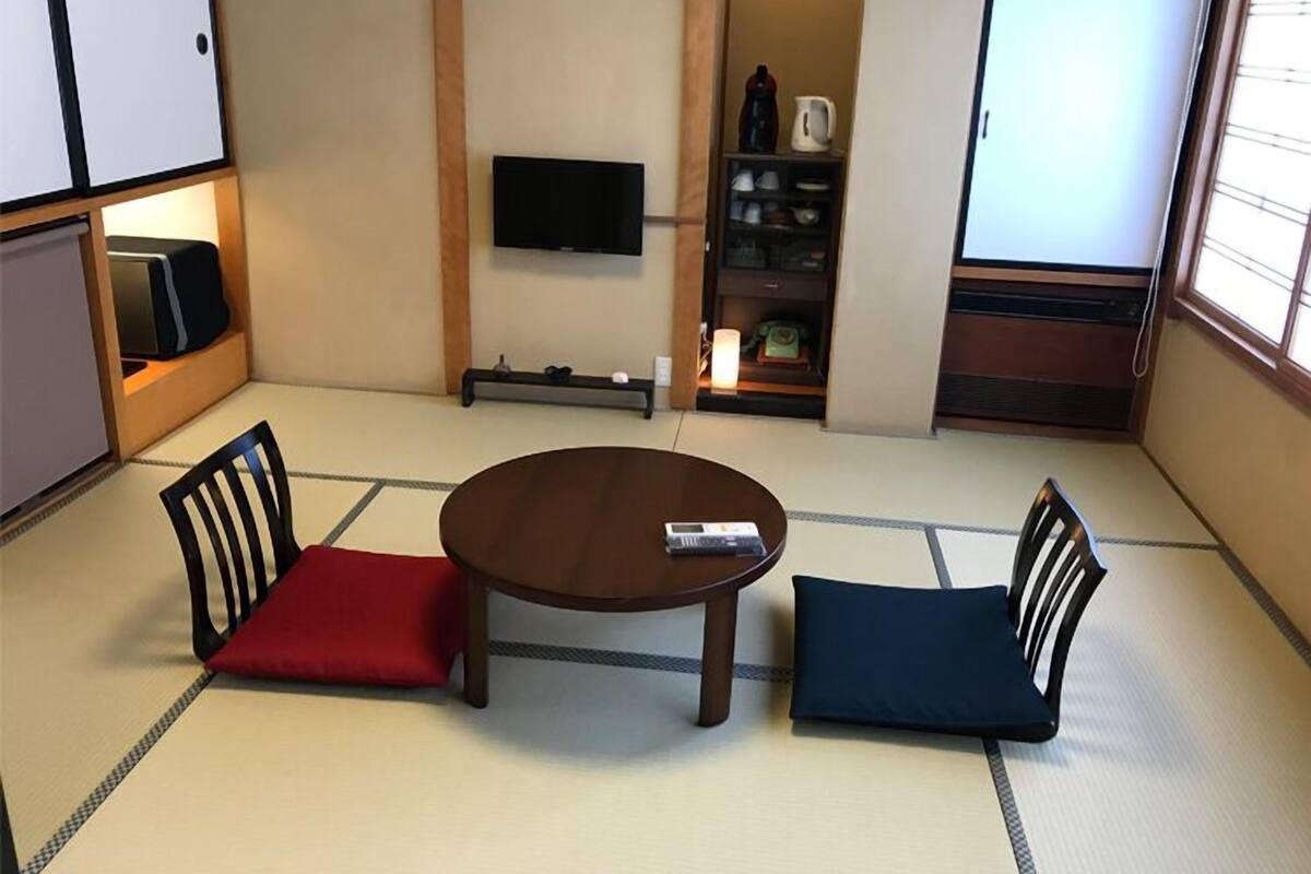Una habitación con tatamis, una mesa baja, dos sillas, un televisor y una estantería con utensilios de cocina.