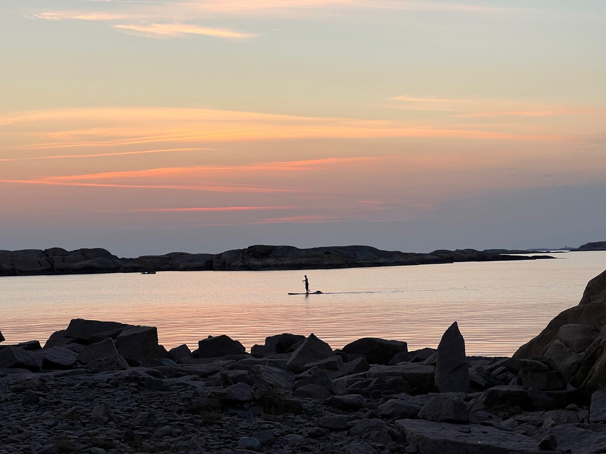 A rocky shoreline at sunset with calm water and a silhouette of a person paddling.