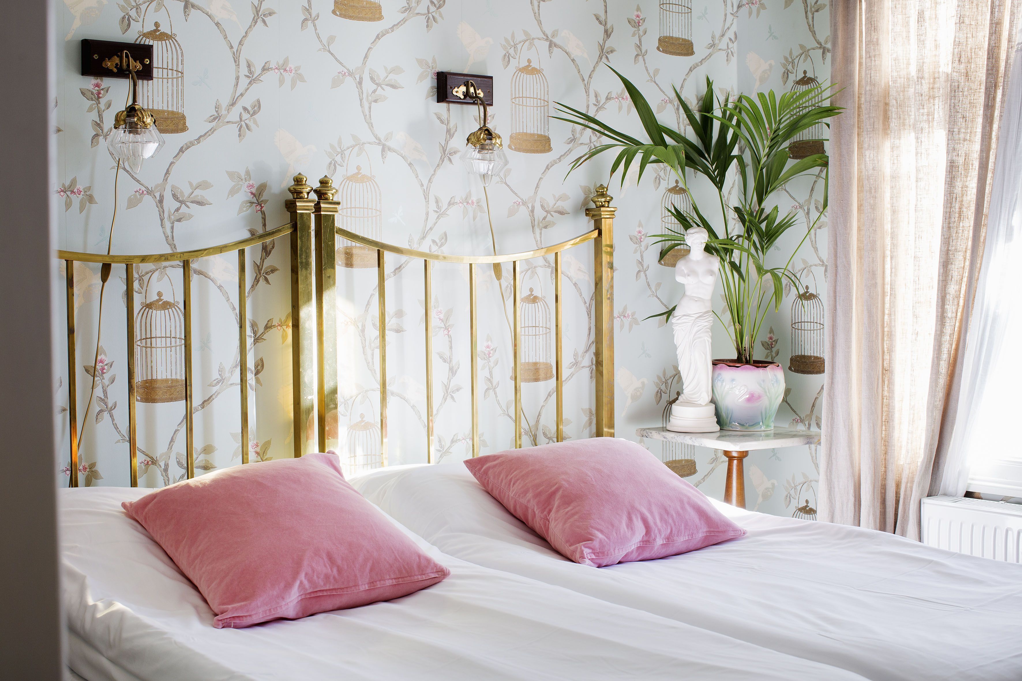 Una cama con ropa de cama blanca y almohadas rosadas, un cabecero con marco dorado, una planta en maceta y una mesita con una estatua.
