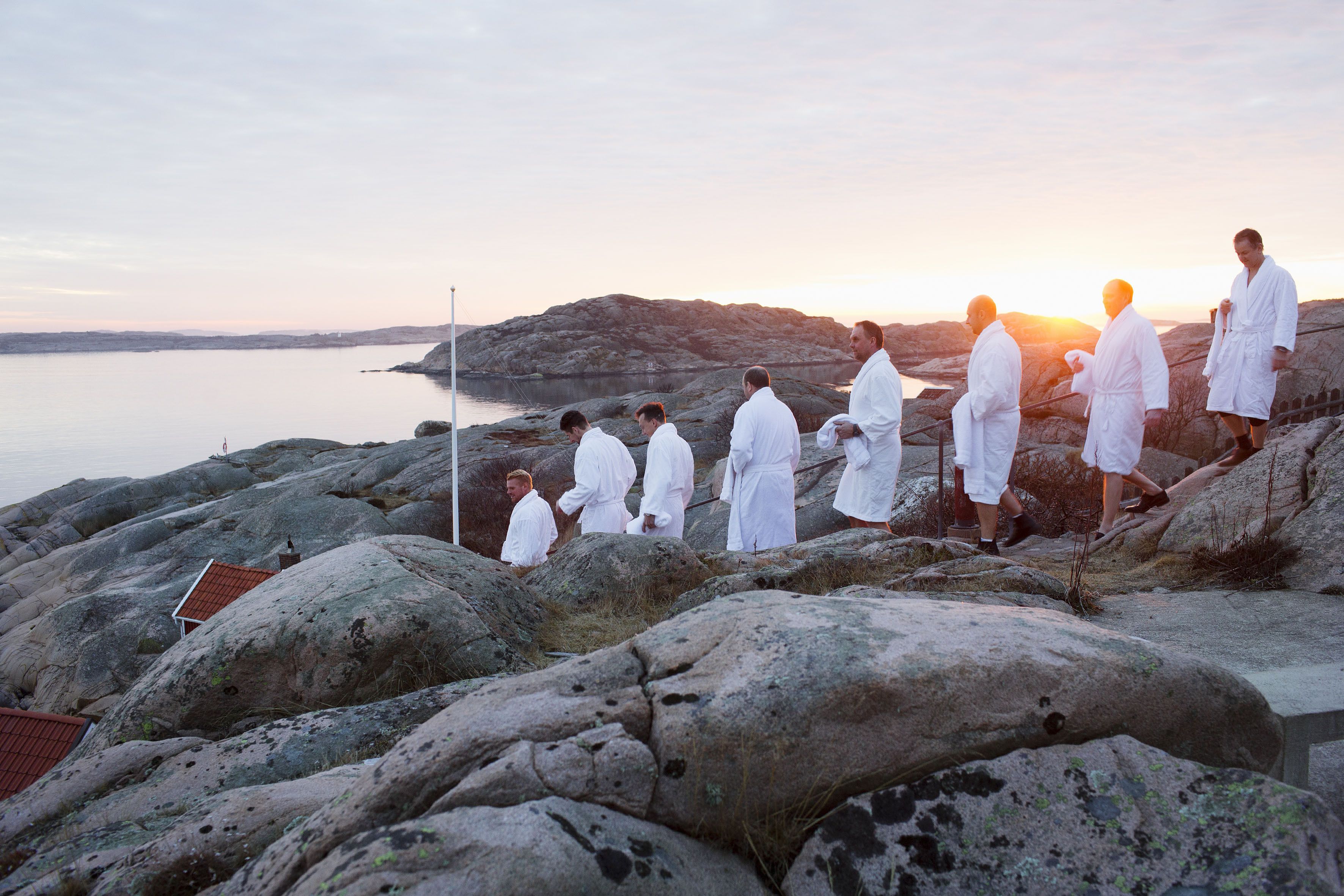 Un groupe de personnes vêtues de robes blanches se tient sur un terrain rocheux, près d’une étendue d’eau, au coucher du soleil.