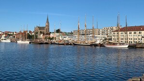 A harbor with several tall sailing ships docked, a prominent church with a tall spire, and a row of modern buildings in the background. - Strandflickornas Havshotell (Lysekil)