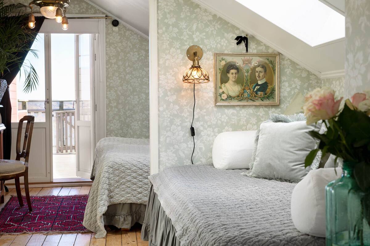 A bedroom with a large window, a chandelier, a framed portrait, and a red rug.