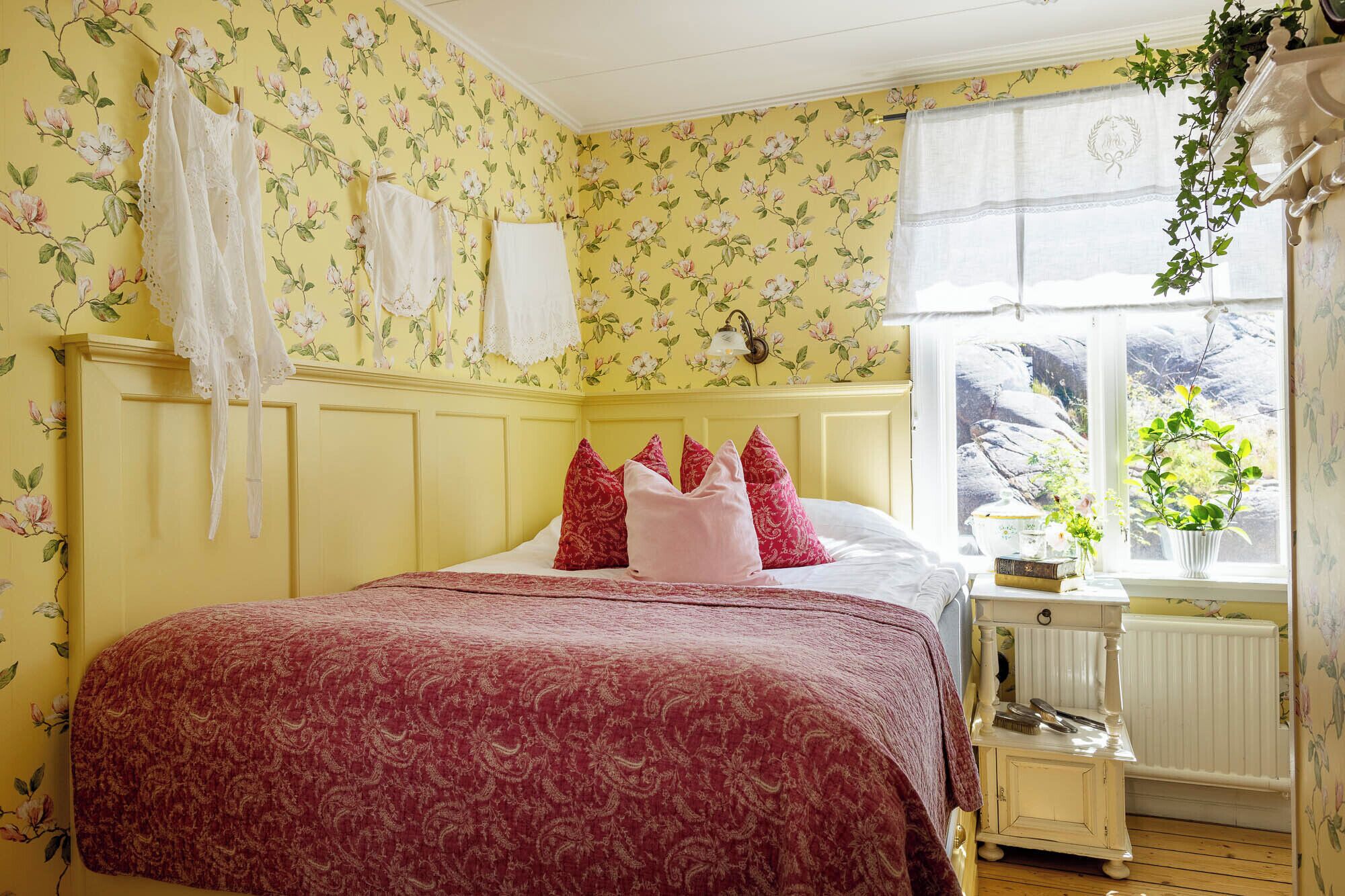 Un dormitorio con papel tapiz floral, una cama con colcha estampada roja y una ventana con vista a rocas.