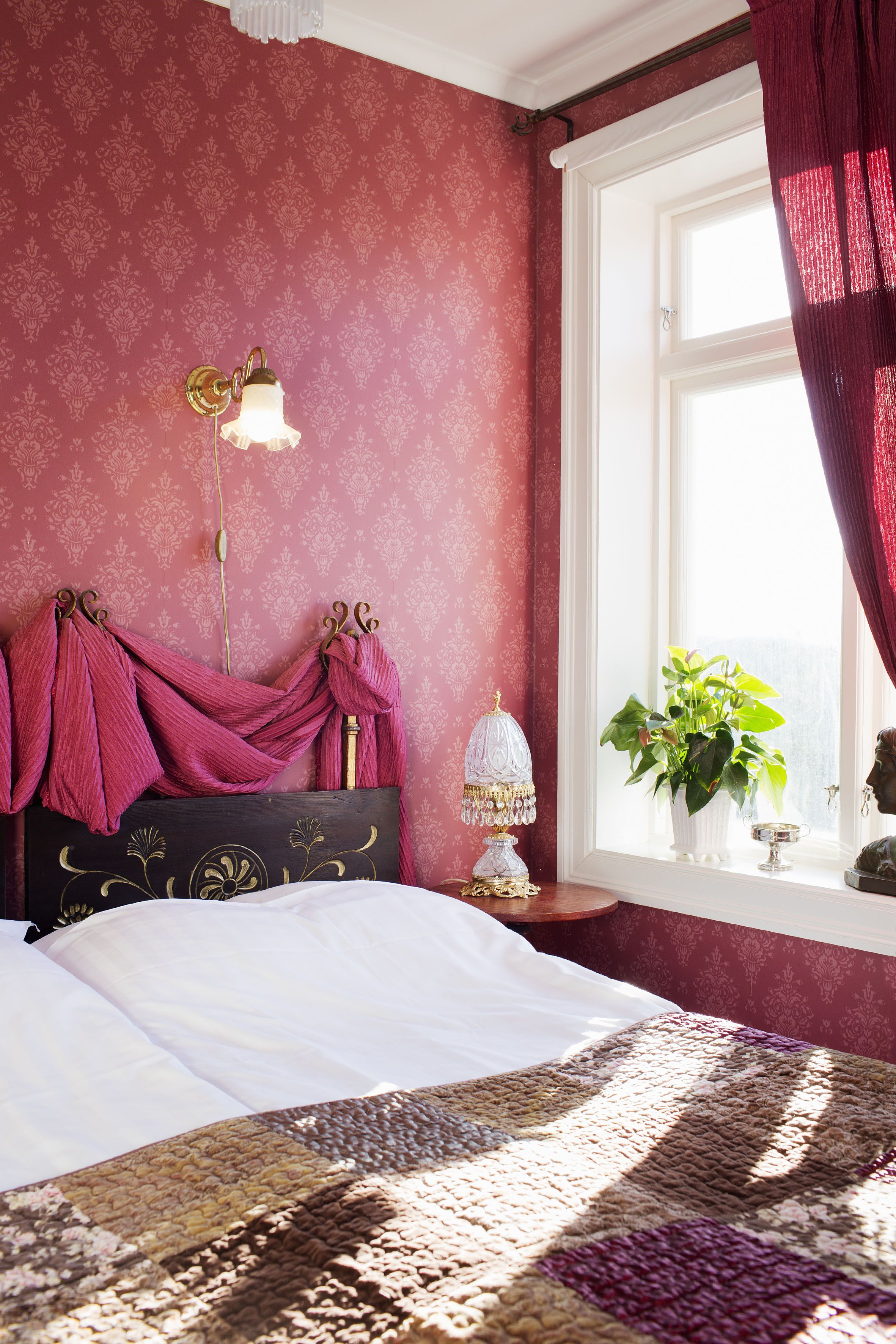 A bedroom with a bed, a nightstand, a lamp, a window with curtains, and a plant.