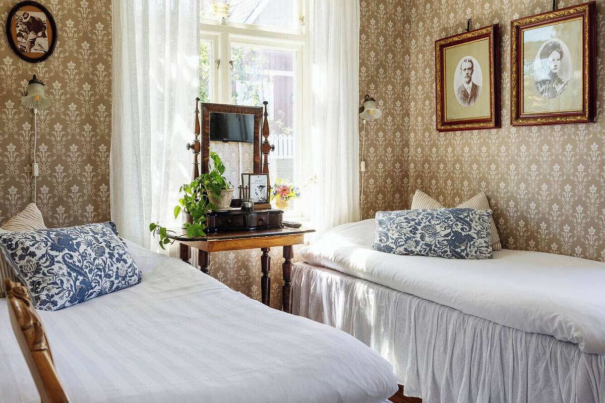 A bedroom with two beds, a wooden nightstand, a desk with a lamp, and two framed pictures on the wall.
