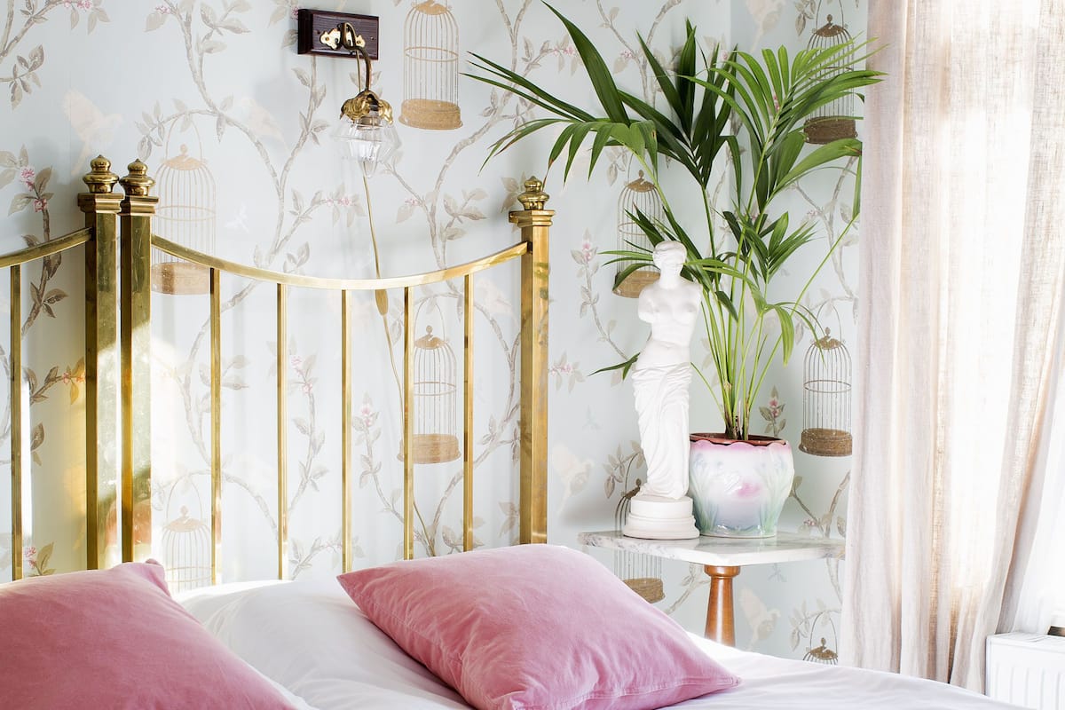 A bedroom with a bed, two pink pillows, a white nightstand, a potted plant, and a wall with a birdcage pattern.