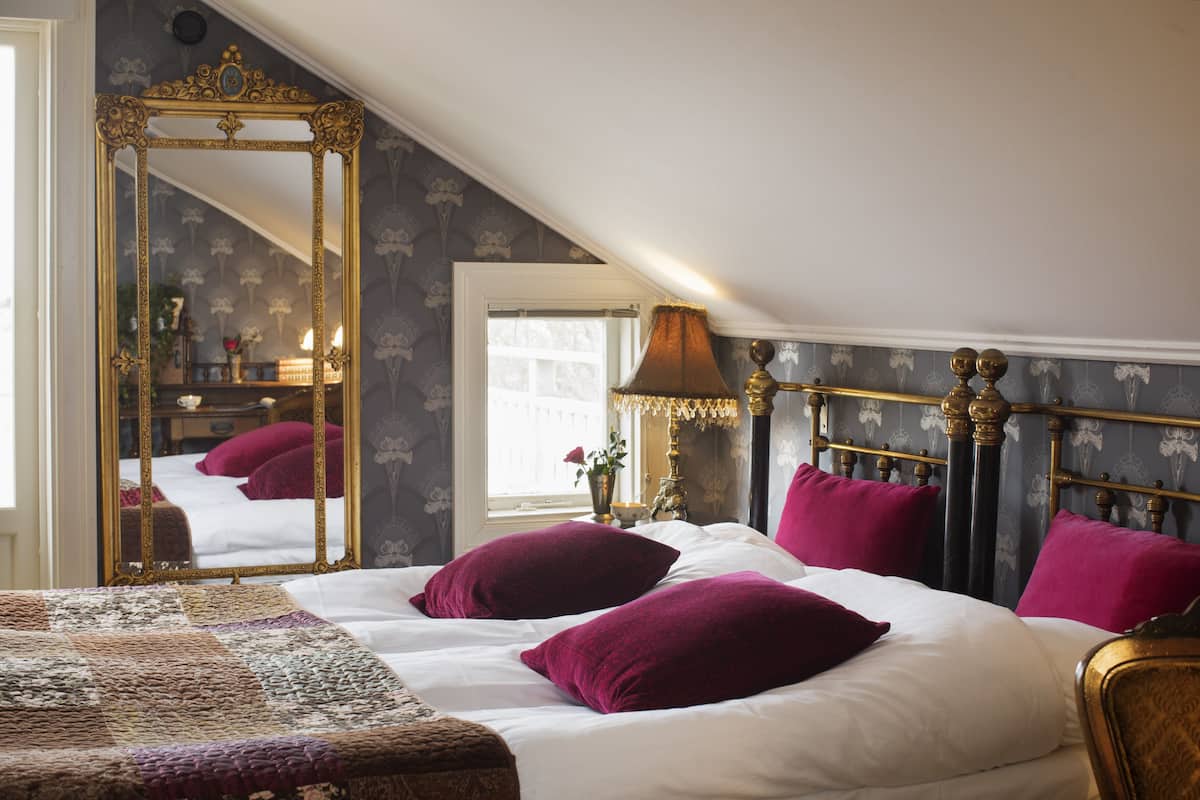 A bedroom with a large ornate mirror, a bed with white linens and purple pillows, a chandelier, and a window with a view of the interior.