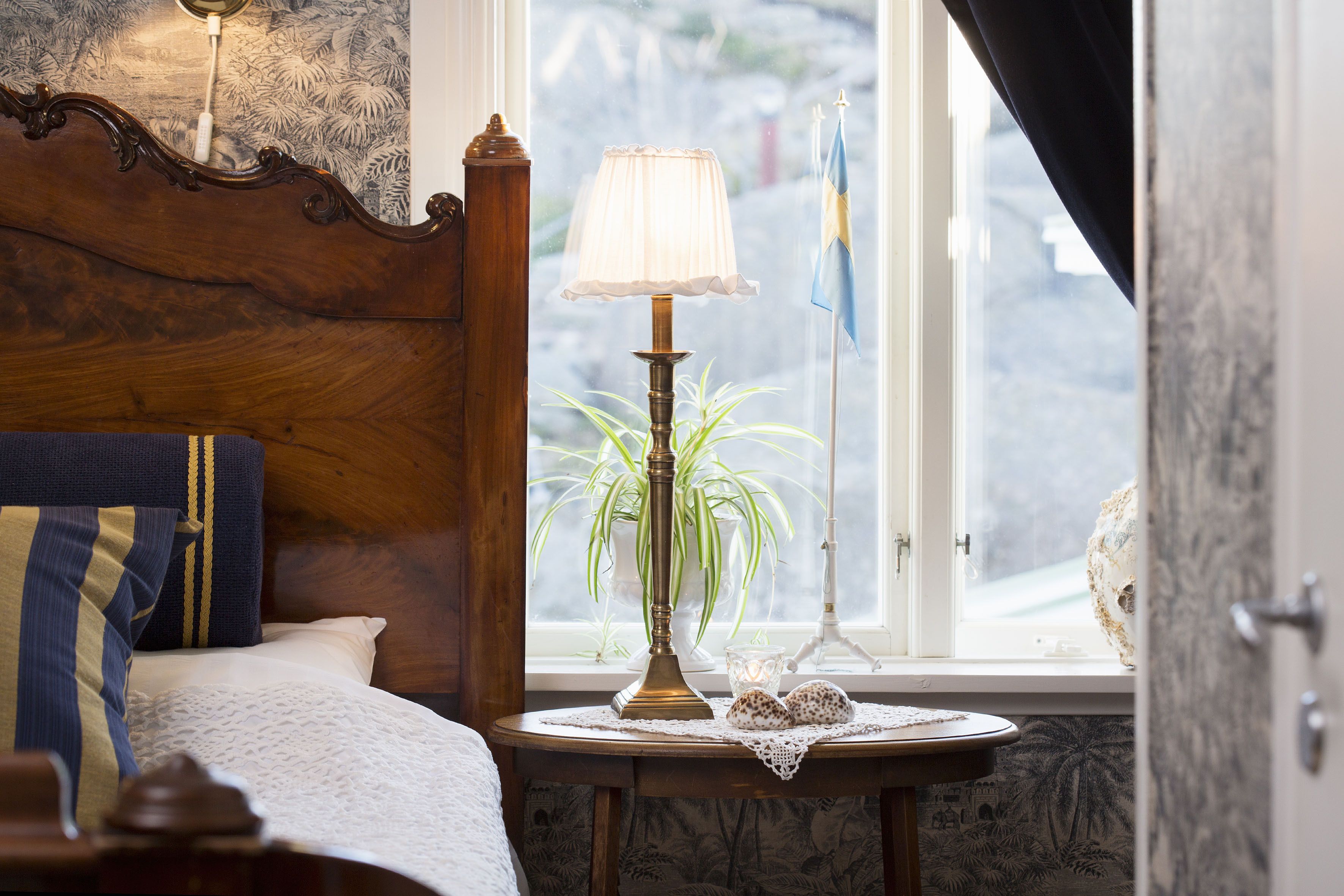 A bedroom with a wooden headboard, a bedside table with a lamp, a plant, and a view of the outside through a window.