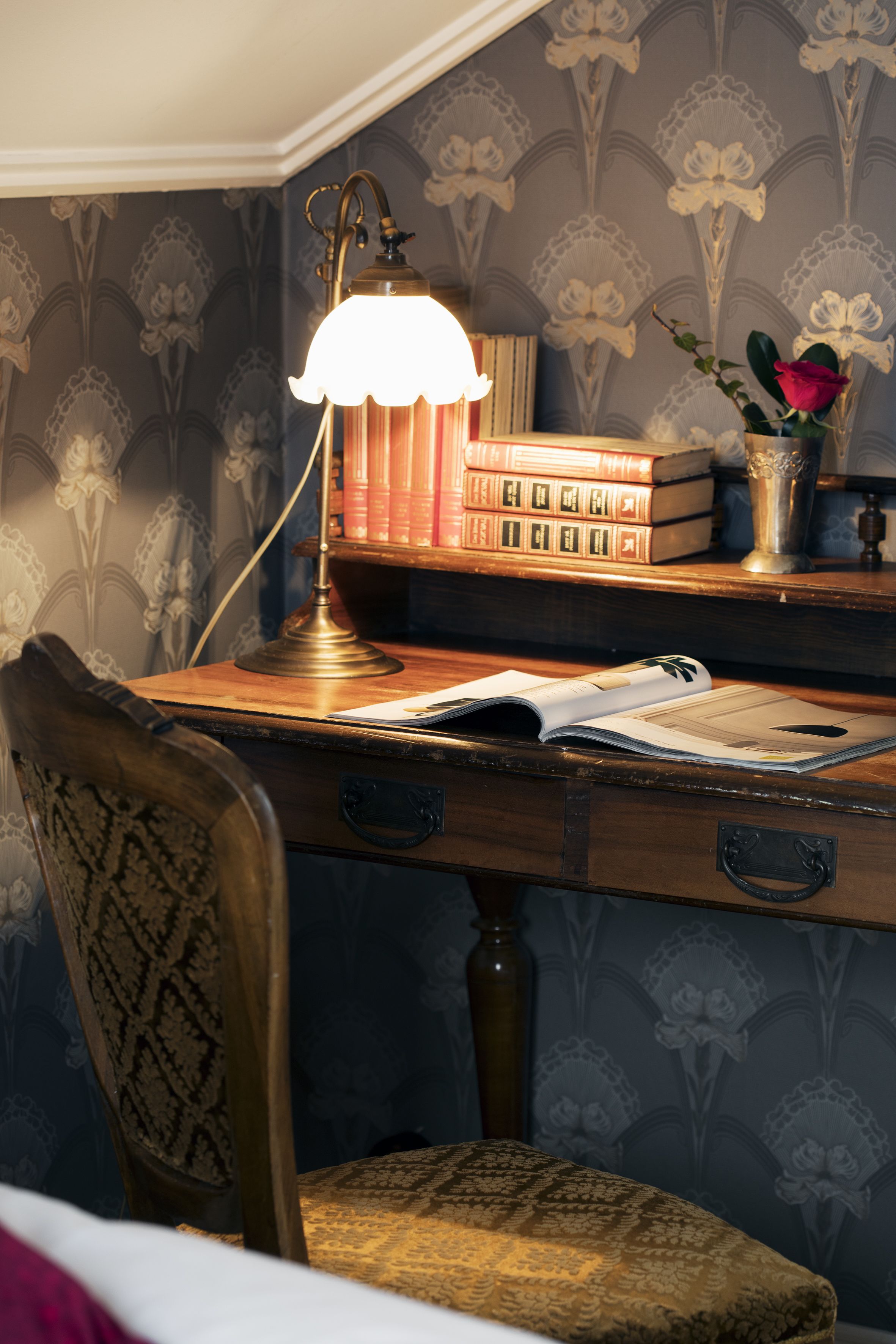A wooden desk with a lamp, books, and a vase with a rose.