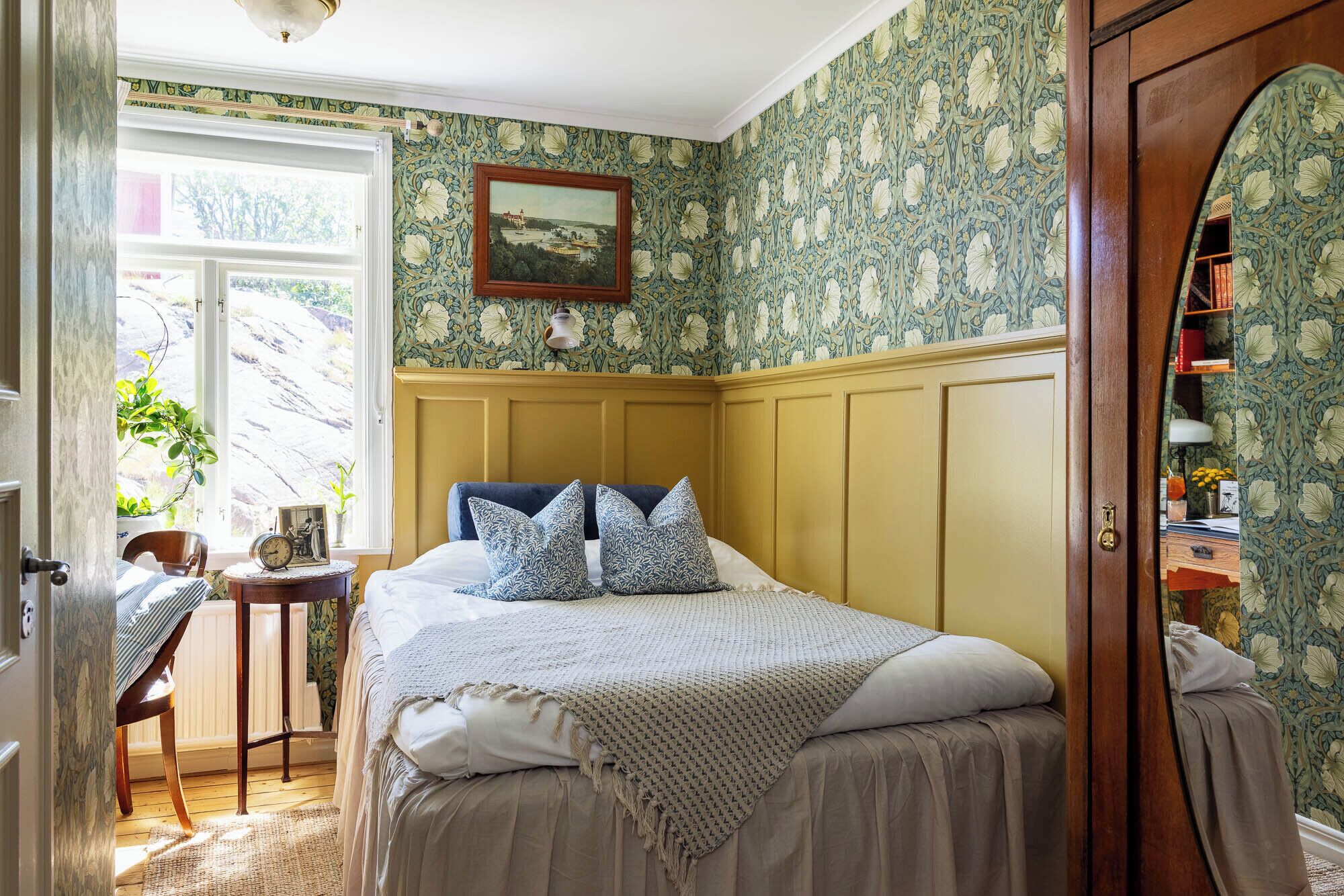 A bedroom with a bed, a wooden headboard, a small table with a clock, and a window with a view of greenery.