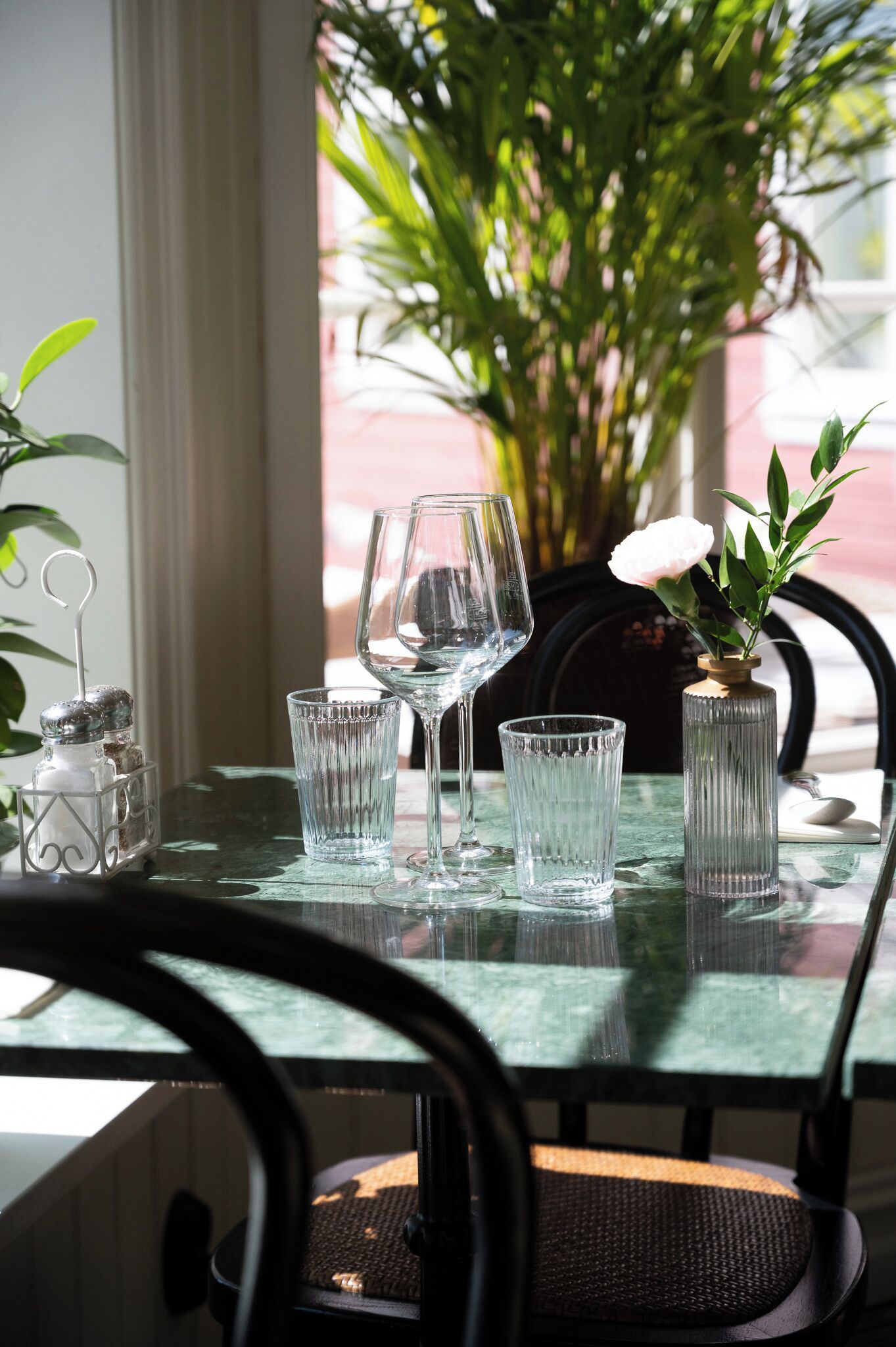 Ett matbord med fyra genomskinliga glas och en vas med en vit blomma, placerat framför ett fönster med utsikt över grönskan.