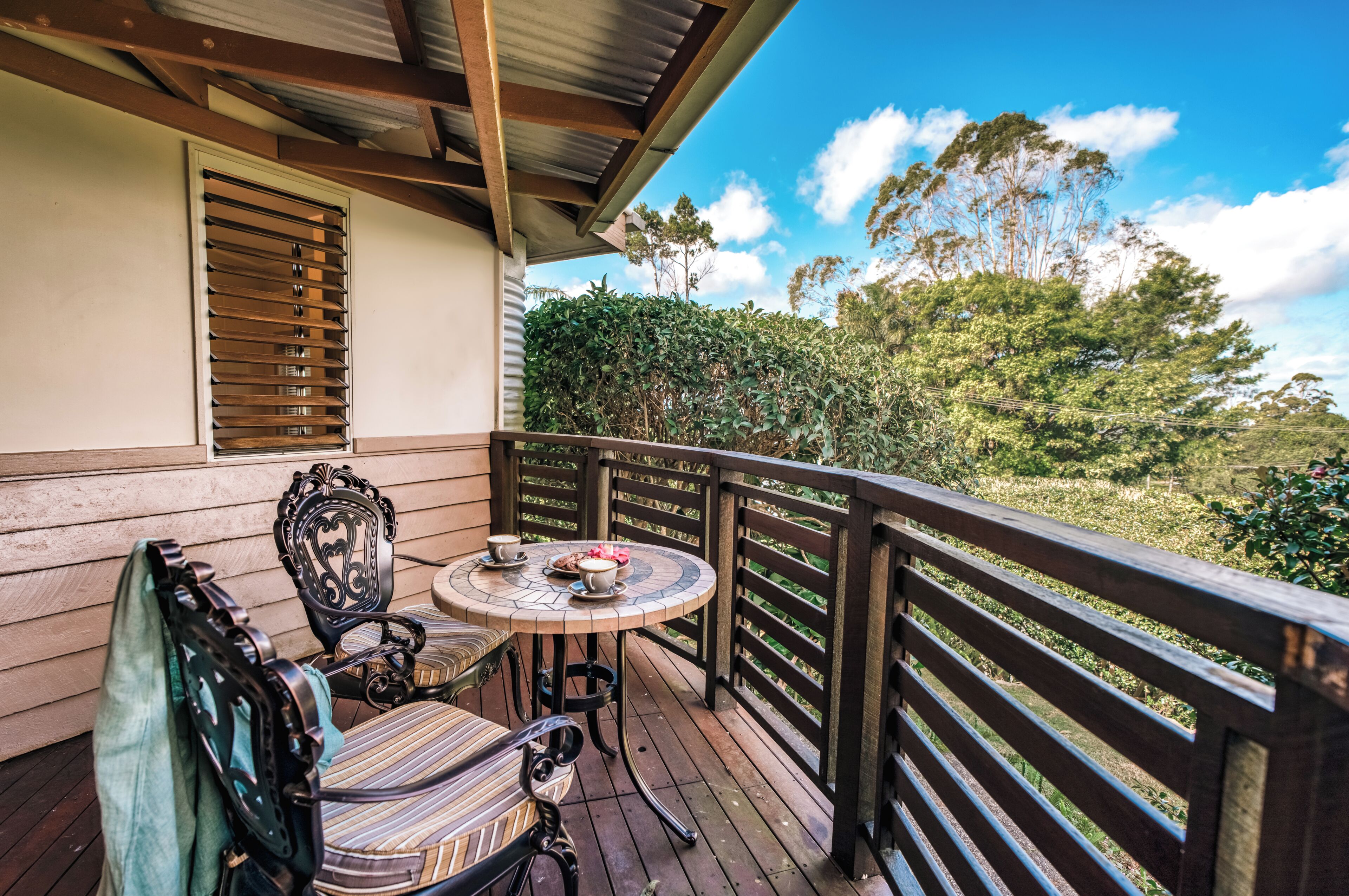 A wooden deck with a round table and chairs, a window with blinds, and a view of trees and sky.