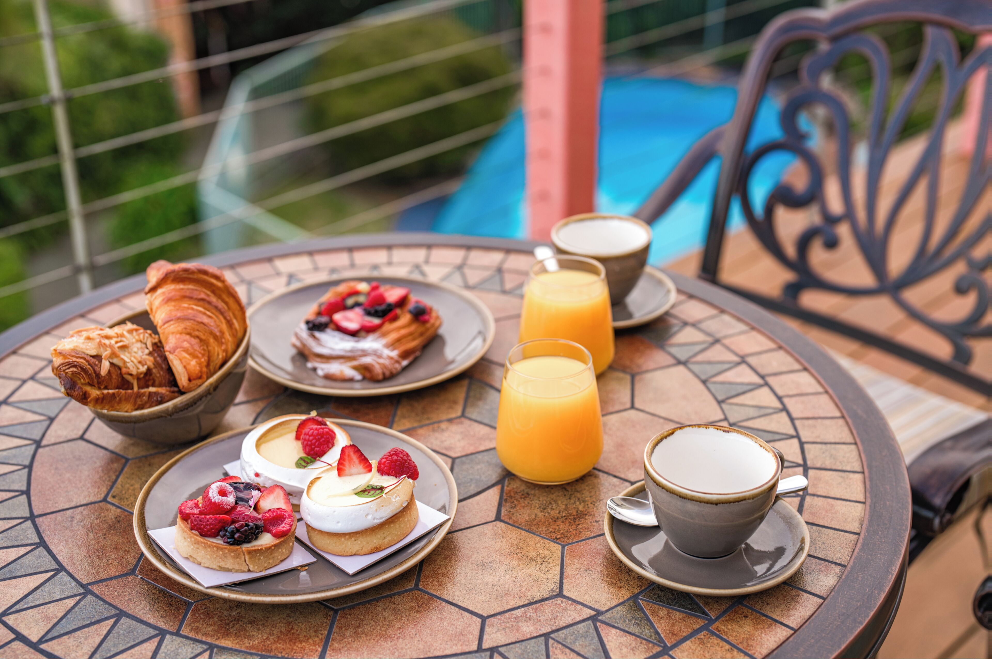 A breakfast setup with croissants, pastries, fruit-topped desserts, orange juice, and coffee on a tiled table with a pool view.