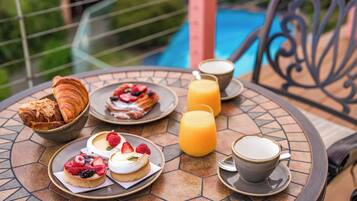 A breakfast setup with croissants, pastries, fruit-topped desserts, orange juice, and coffee on a tiled table with a pool view.