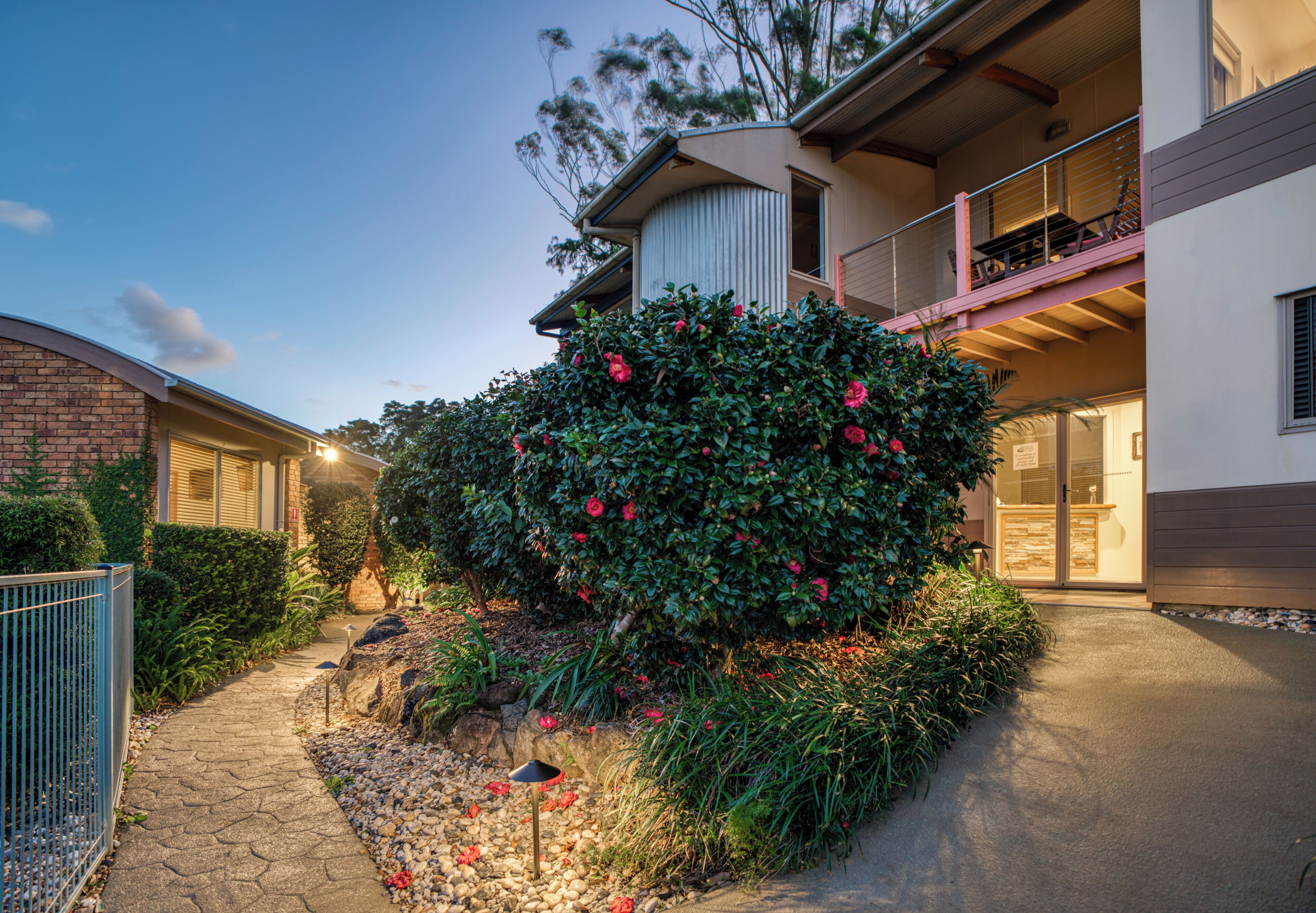 A modern two-story building with a balcony, a well-maintained garden, and a pathway leading to the entrance.