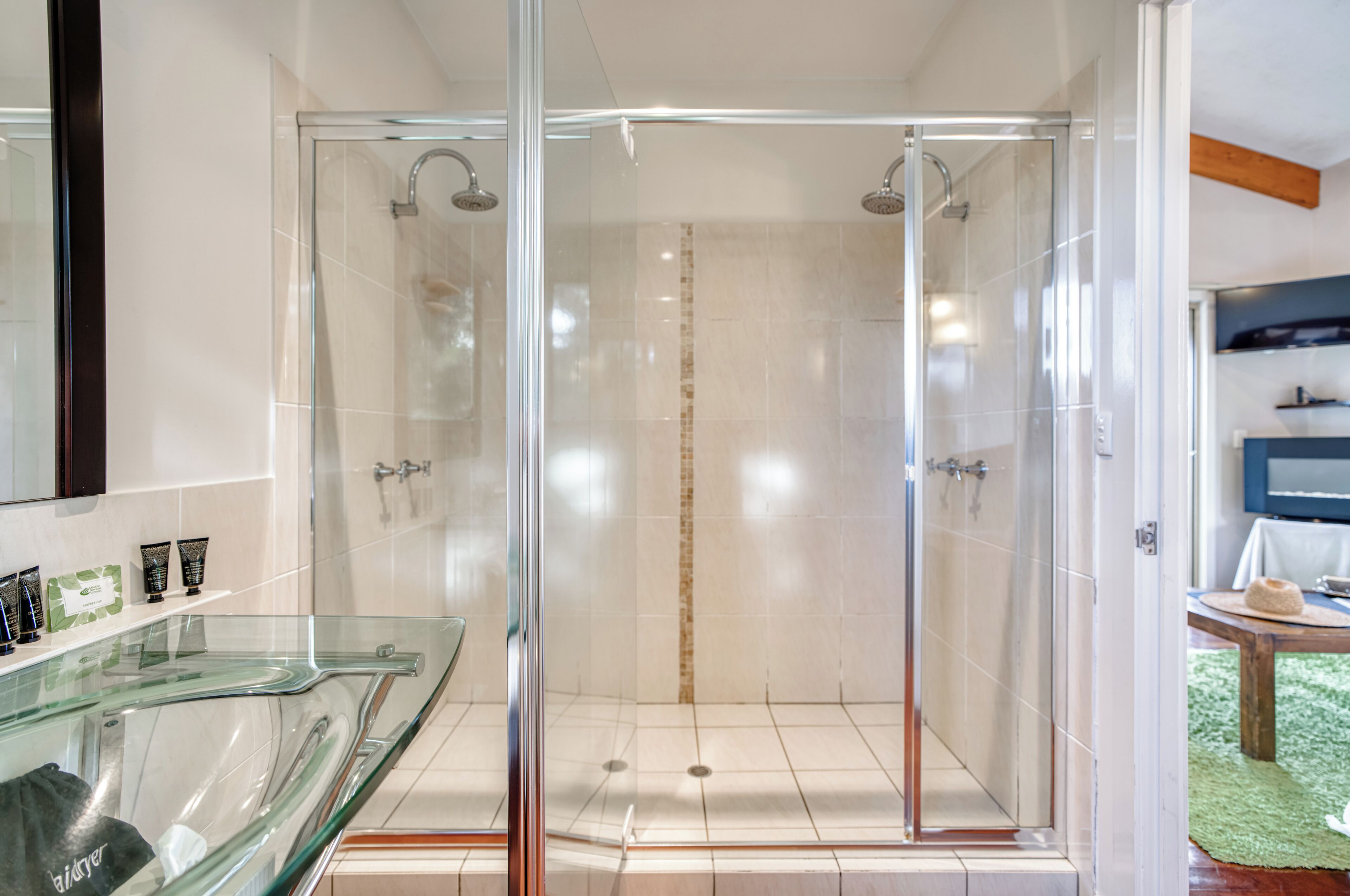 A glass-enclosed shower area with a glass partition, a glass countertop with toiletries, and a wooden ceiling beam.
