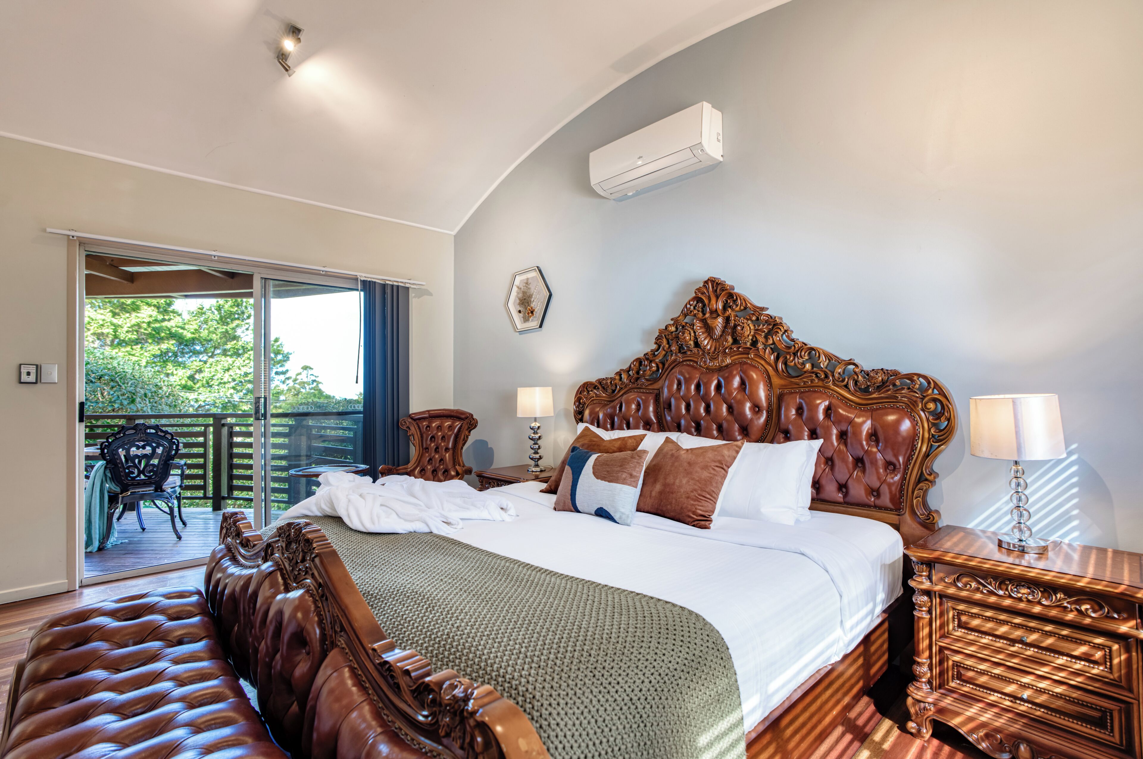 A bedroom with a large bed, a wooden headboard, a nightstand, a lamp, and a view of the outdoors.
