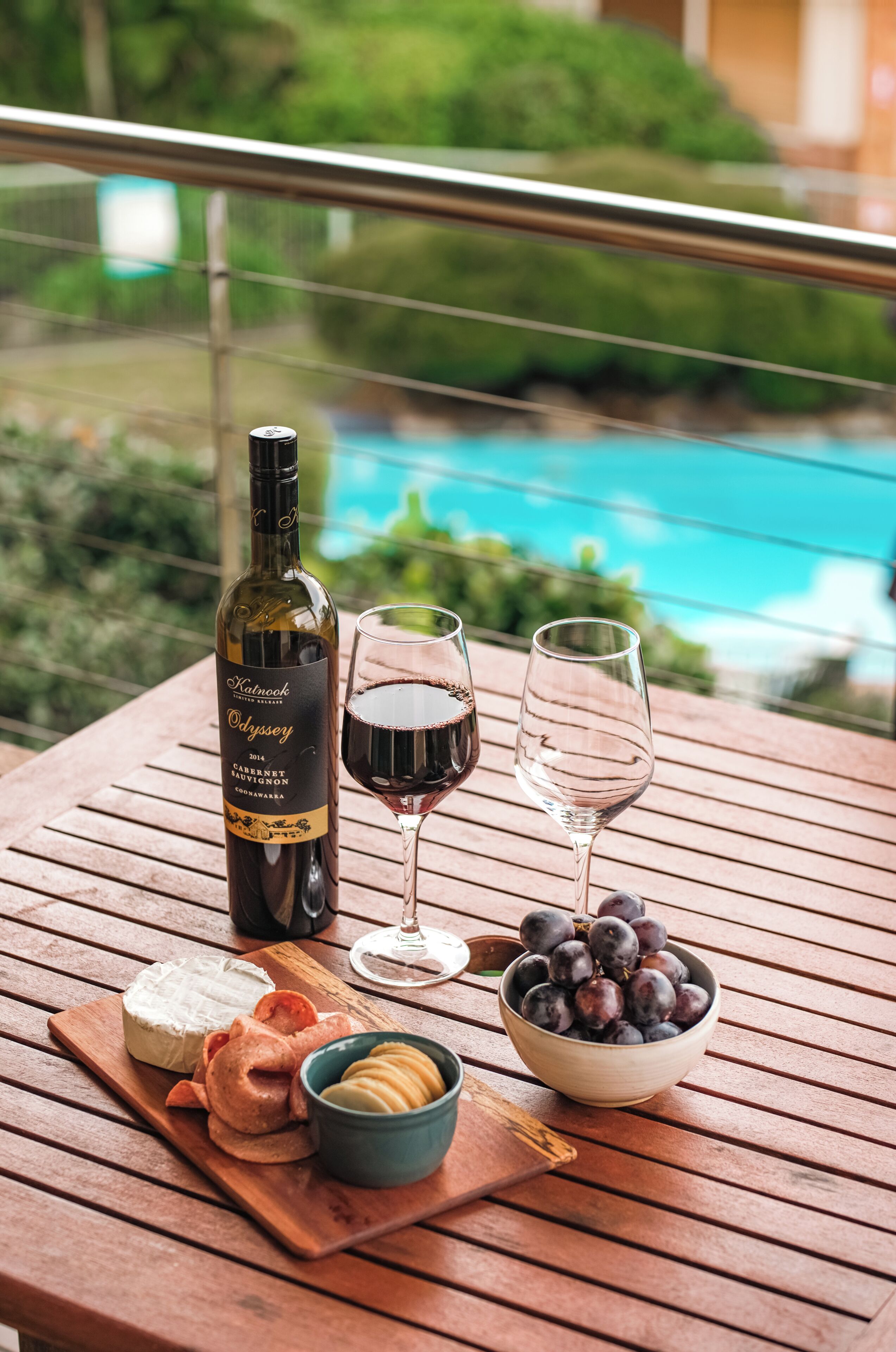 A wooden table with a bottle of wine, two glasses, a bowl of grapes, a bowl of crackers, and a piece of cheese.