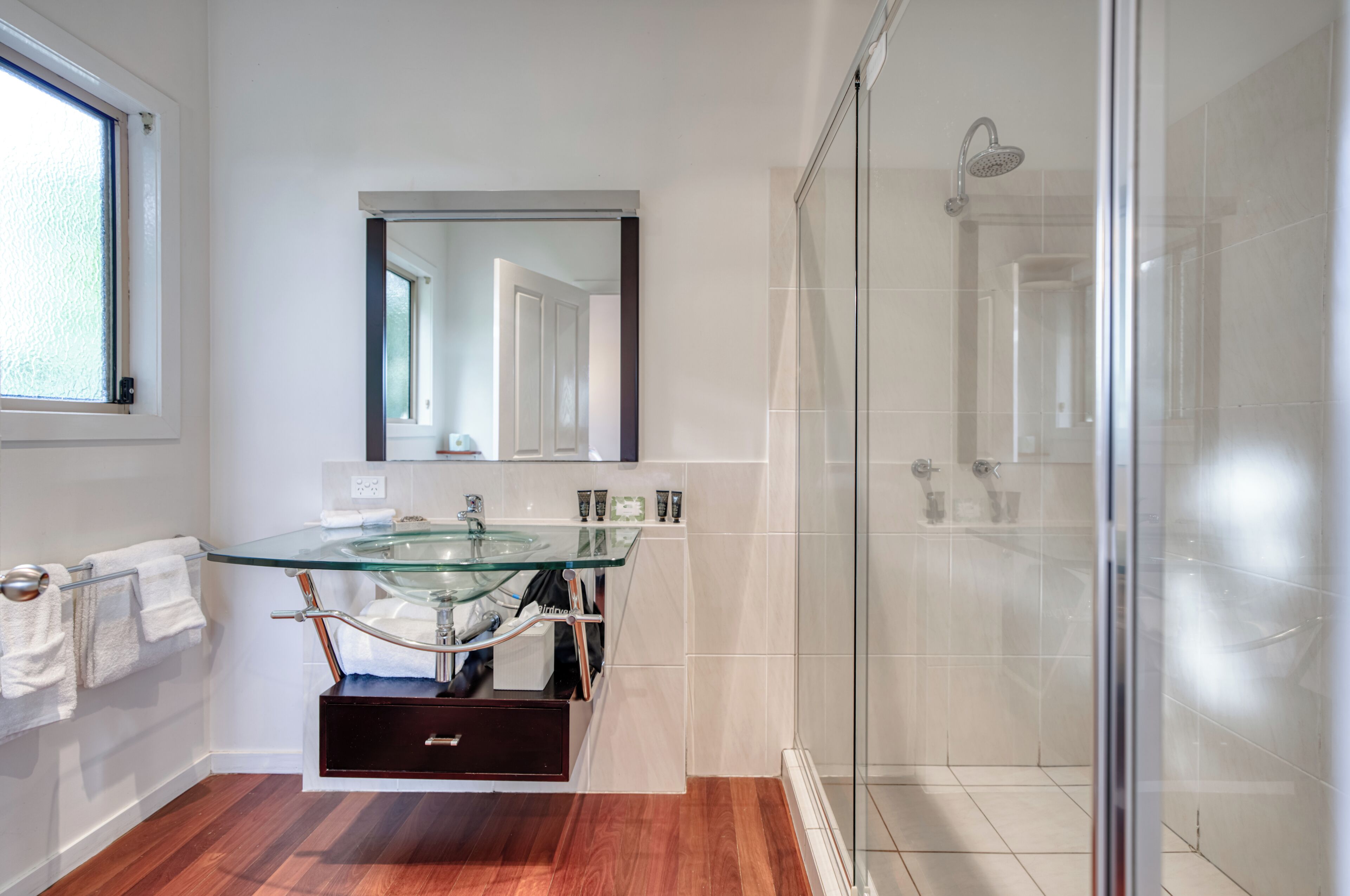 A modern bathroom with a glass sink, a large mirror, a glass-enclosed shower, and wooden flooring.