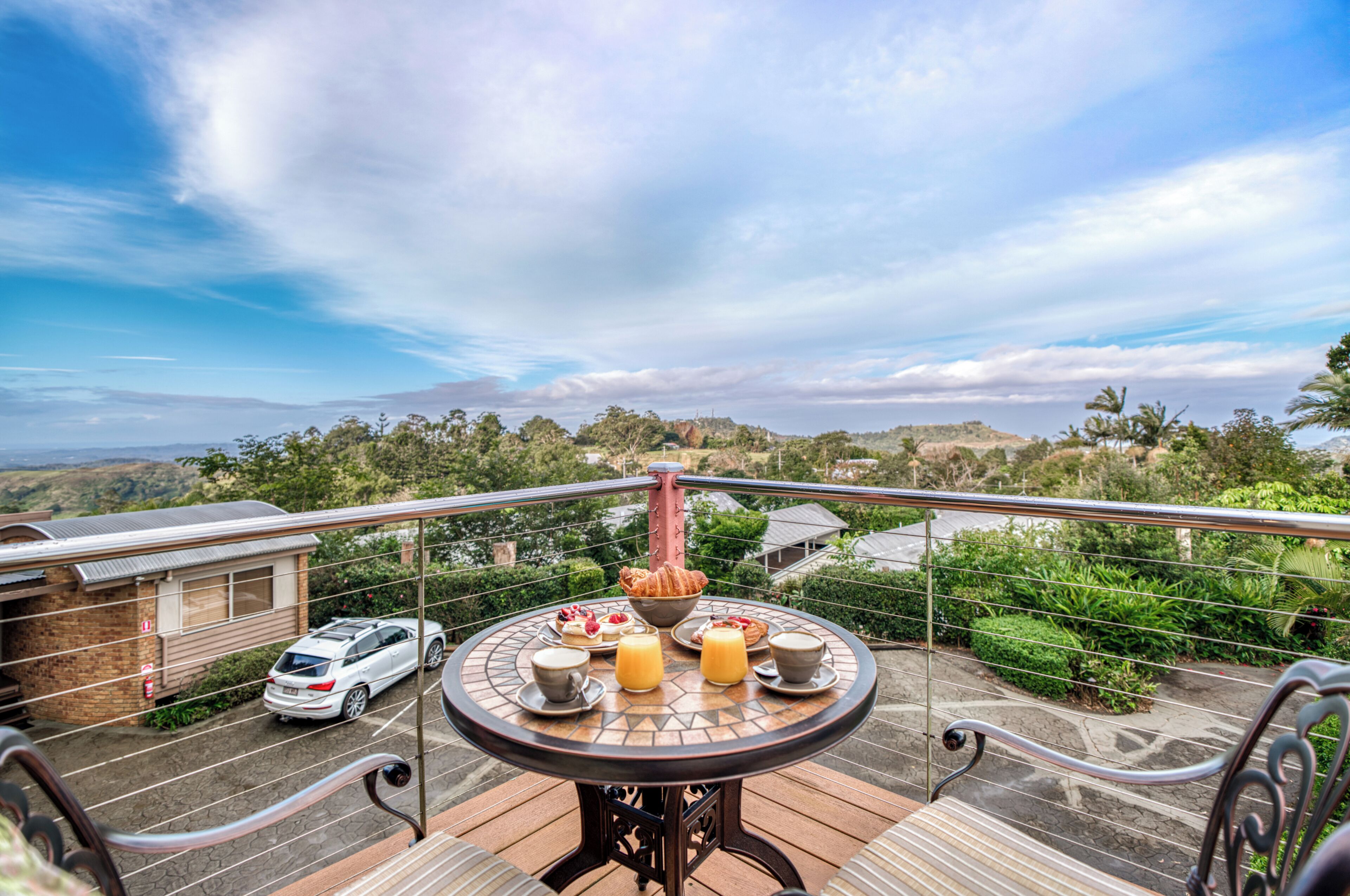 A balcony with a table set for breakfast, overlooking a parking area and a scenic landscape.