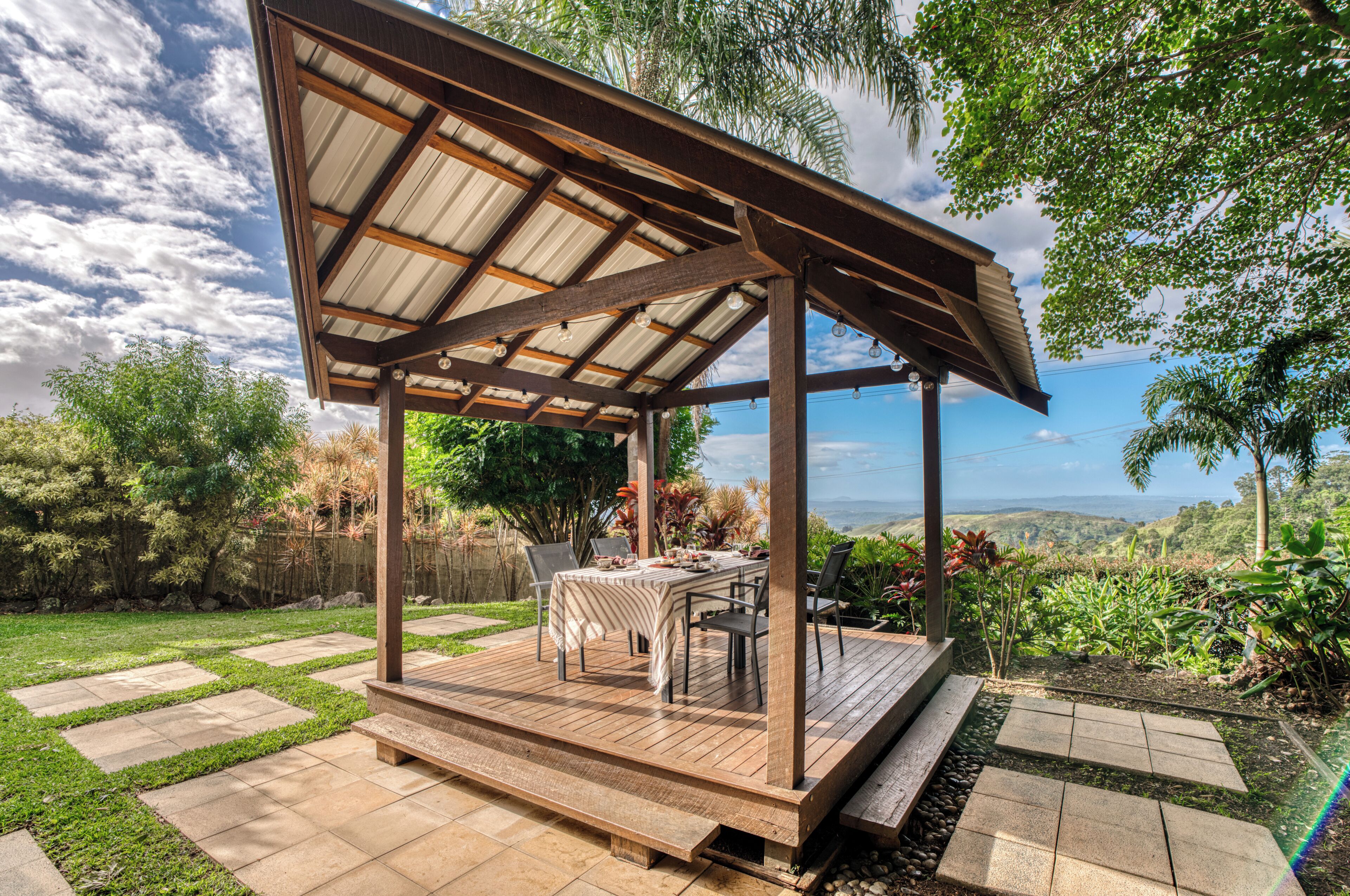 A covered outdoor patio with a table and chairs, surrounded by a garden and a view of the landscape.