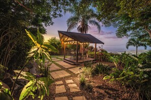 A covered outdoor seating area with a table and chairs, surrounded by a garden with various plants and a stone pathway. - Maleny Terrace Cottages (Maleny)