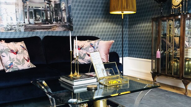 A living room with a dark blue patterned wall, a black sofa with floral cushions, a glass coffee table with books and candles, a gold lamp, and a wooden cabinet with glass doors. - Annas Hotell (Kristianstad)